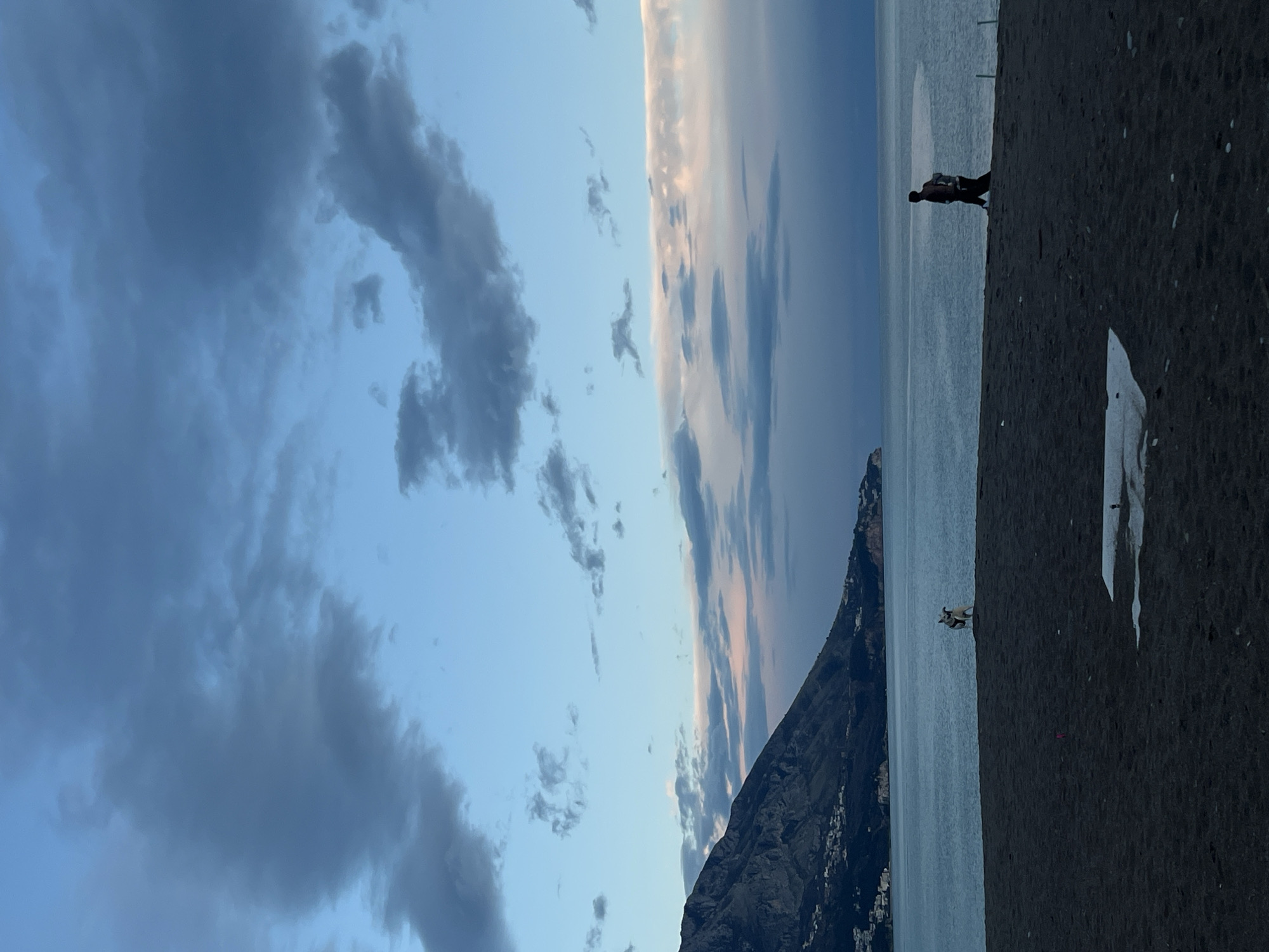 A horizontal outdoor shot depicts a coastal scene in Bečići, Montenegro, during twilight. The sky, covering the left half of the frame, shows various shades of blue with darker gray clouds on the far left and lighter, near-white streaks towards the middle. To the right of the sky, a body of water extends towards the horizon. A mountain range with dark, rocky textures is visible along the water's edge. The right side of the image is dominated by dark, coarse sand, possibly a beach. Two human figures are present on the sand: one appears to be walking along the shore near the water's edge, and another is slightly further away, with a smaller animal, likely a dog, standing near it. The lighting suggests either sunrise or sunset.