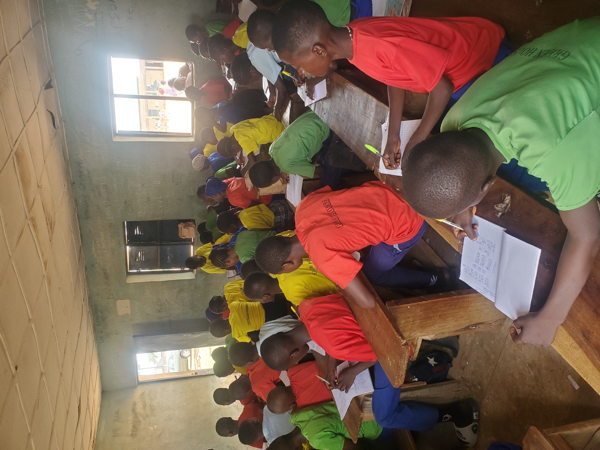 Inside a classroom in Akwanga, Nigeria, numerous children are seated at wooden desks. They are dressed in red, yellow, and green shirts and are writing on papers. Sunlight enters through windows in the room. The classroom walls are light green and have writing on them.