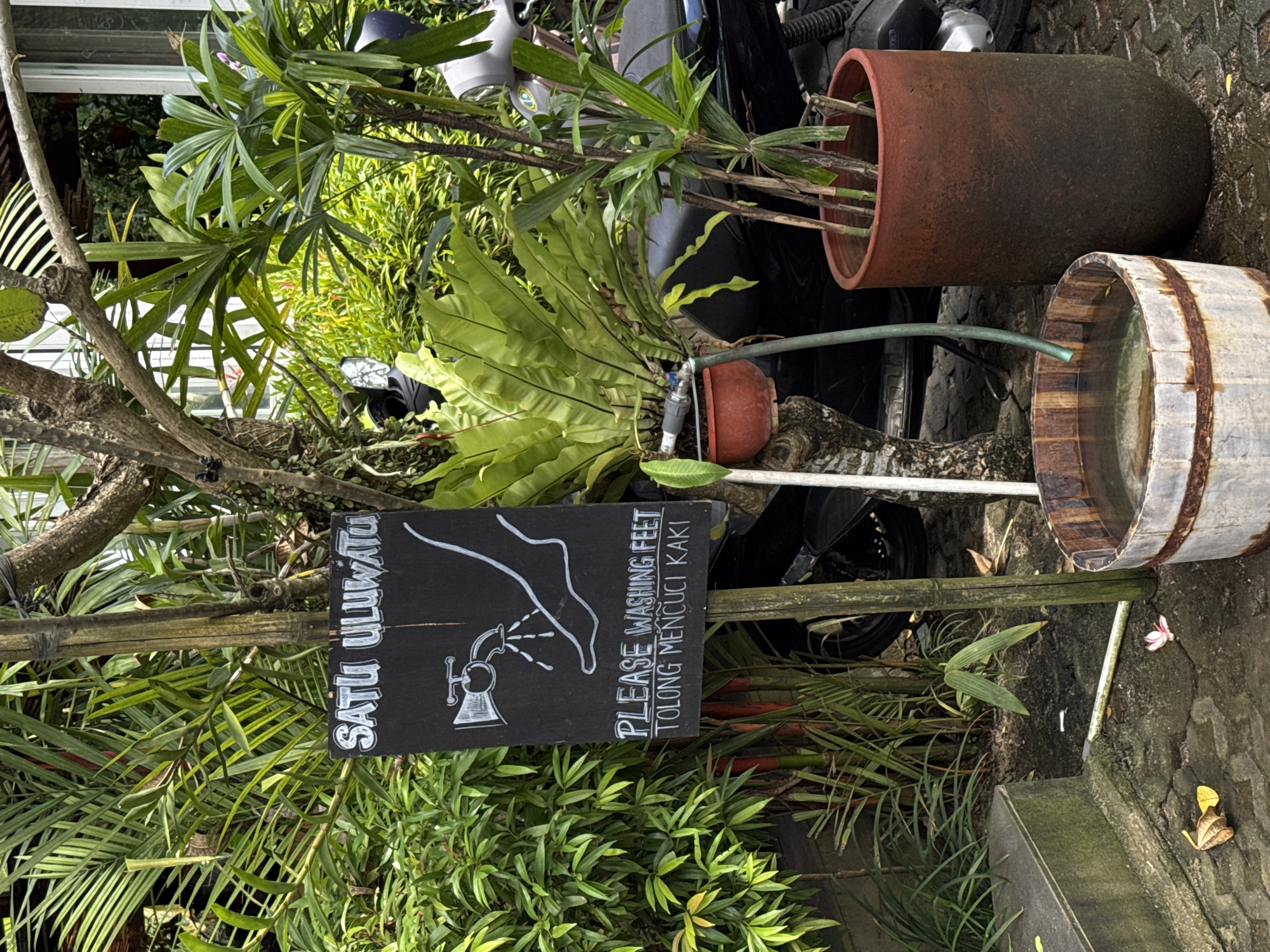 The image shows a foot-washing station in Kuta Selatan, Indonesia. A black sign with white lettering reads "SATU ULUWATU" and "PLEASE WASHING FEET TOLONG MENCUCI KAKI," accompanied by a drawing of a foot under a faucet. The sign is mounted on a bamboo pole, surrounded by various plants. To the right, there is a wooden barrel filled with water. Behind the barrel, there are terracotta pots containing plants and a black motorcycle. The ground is stone. The scene appears to be outdoors.