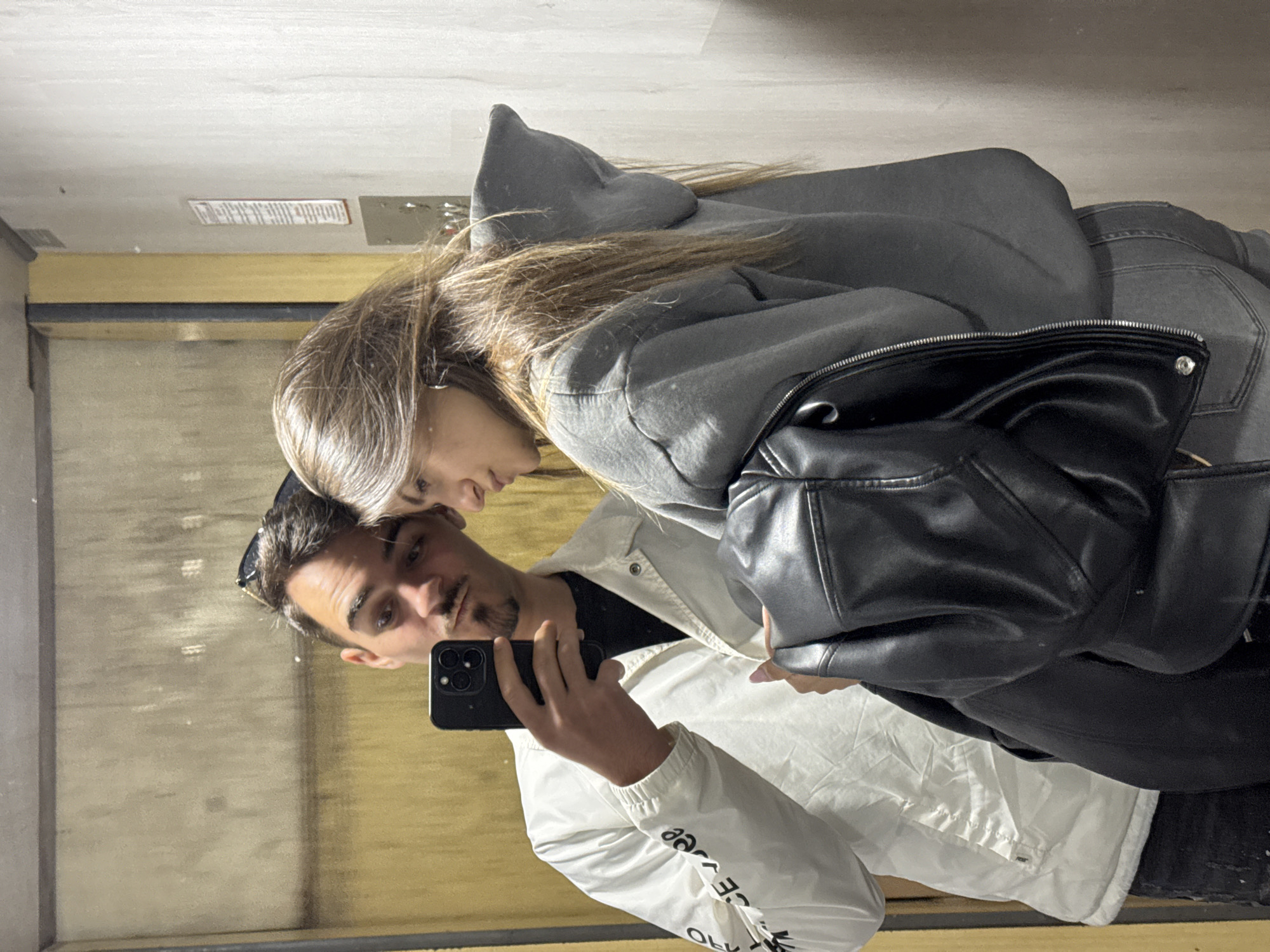An indoor mirror selfie captures a man and a woman in what appears to be an elevator or a narrow hallway with reflective walls. The image is presented vertically, rotated 90 degrees counter-clockwise.
The man, positioned in the foreground, is holding a black smartphone with a triple camera lens, presumably taking the photo. He has dark hair, a mustache, and a short beard (goatee), with sunglasses resting on his head. He is wearing a black t-shirt and a white lightweight jacket. Partially visible and reversed text on his jacket sleeve includes letters like 'ee', 'Y', and 'OLK'.
Behind and slightly to the left of the man, the woman leans in close, looking downwards towards the phone or the man. She has long, light brown hair. She is dressed in a grey hooded sweatshirt, grey jeans, and a black leather jacket worn over the hoodie.
The background visible in the reflection consists of light-colored walls or ceiling, with a visible rectangular sign or vent on the upper wall whose text is unreadable due to distance and focus. A wooden or metal panel runs horizontally across the mid-section of the reflection. The overall scene suggests a casual moment between the two individuals. The location context provided is Sofia, Bulgaria, although no specific visual cues within the image confirm this.