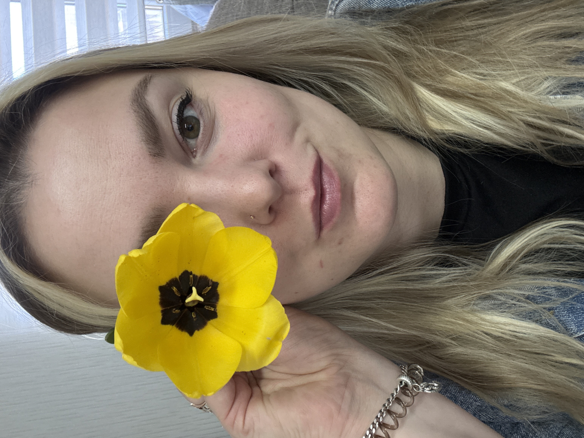 A fair-skinned woman is holding a yellow tulip in front of her face. She has blonde hair, and is wearing a nose ring and a silver charm bracelet. She appears to be wearing a denim jacket. The background is blurred, but shows a window with vertical blinds. There is no information in the image that would suggest a location in the city of Noviy, Russia.