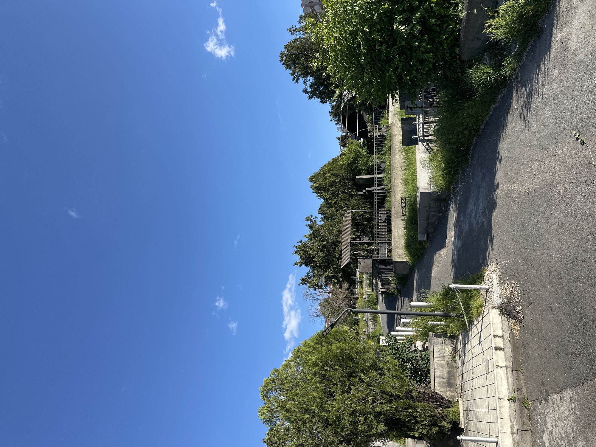 The image depicts a section of a cemetery in Yerevan, Armenia. A paved pathway runs along the edge of the cemetery, bordered by grave markers and vegetation. The sky is blue with scattered clouds. There are no people visible in the frame. Trees and shrubs are present both within the cemetery and along its perimeter. A metal fence separates the cemetery from the pathway. A few streetlamps and a chain link fence line the walkway. The road surface appears slightly worn.