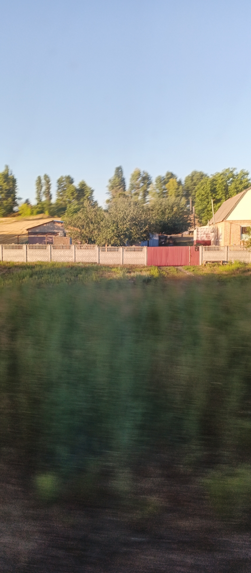 The image displays a rural scene with a distinct linear blur, suggesting motion. In the midground, a low, grey concrete fence runs horizontally across the frame. Behind the fence, there is a red metal gate. Further back, a light-colored brick house with a sloped roof is visible on the right. To the left of the house, there are several trees and structures, including a building with a corrugated metal roof and a section of what appears to be a harvested field, possibly containing hay. The background is dominated by a clear, light blue sky. The foreground is heavily blurred with horizontal streaks of dark green and black, indicating a fast-moving perspective, likely from a vehicle.
