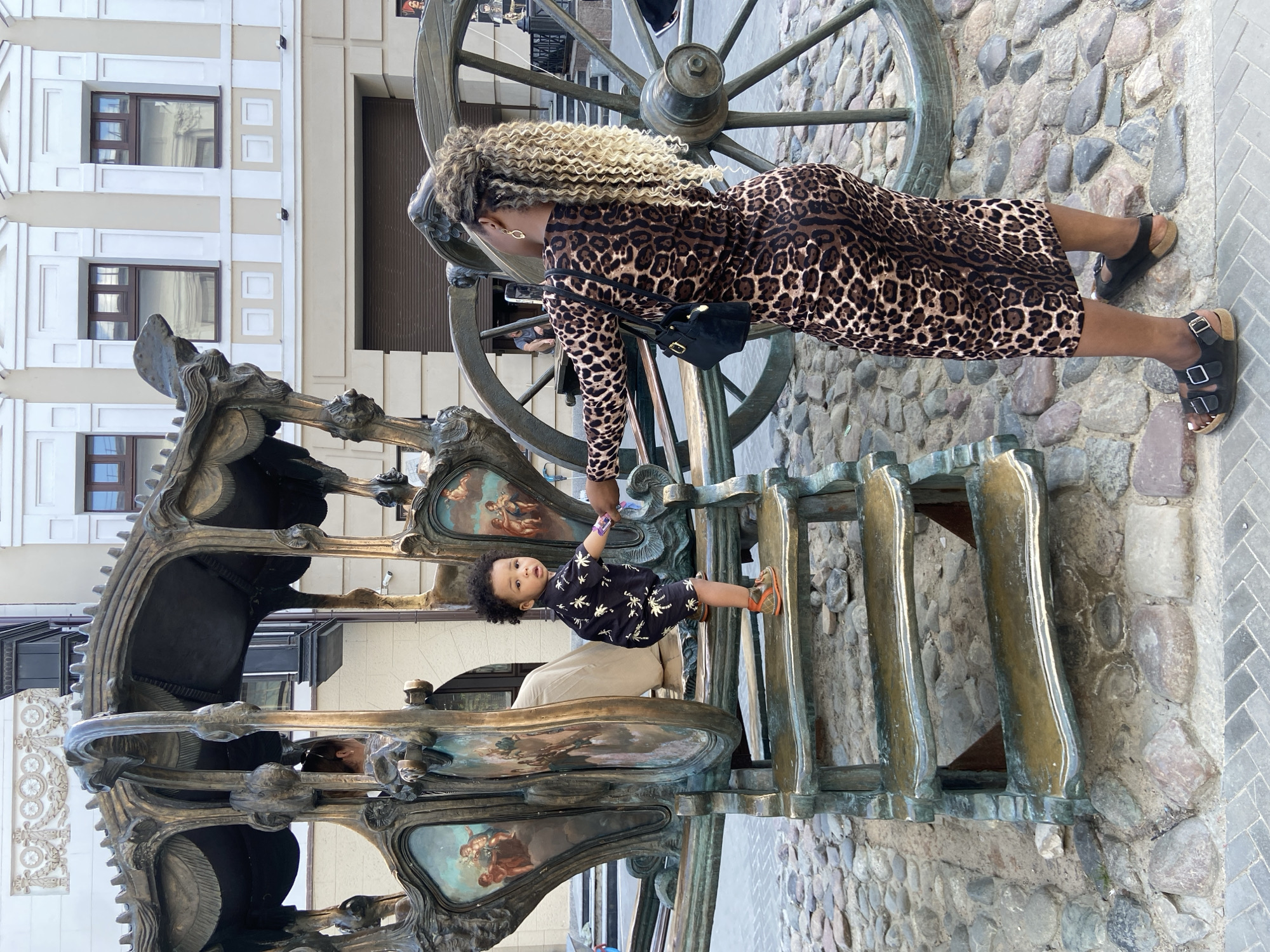 A woman in a leopard-print dress and black sandals is standing in front of a large, bronze monument of a carriage. She is reaching down to hold the hand of a young child in a black shirt with white designs and orange shoes, who is standing on the carriage. The monument is located on a stone path, with a building visible in the background. The location is likely Kazan, Russia, based on the style of the monument and building.
