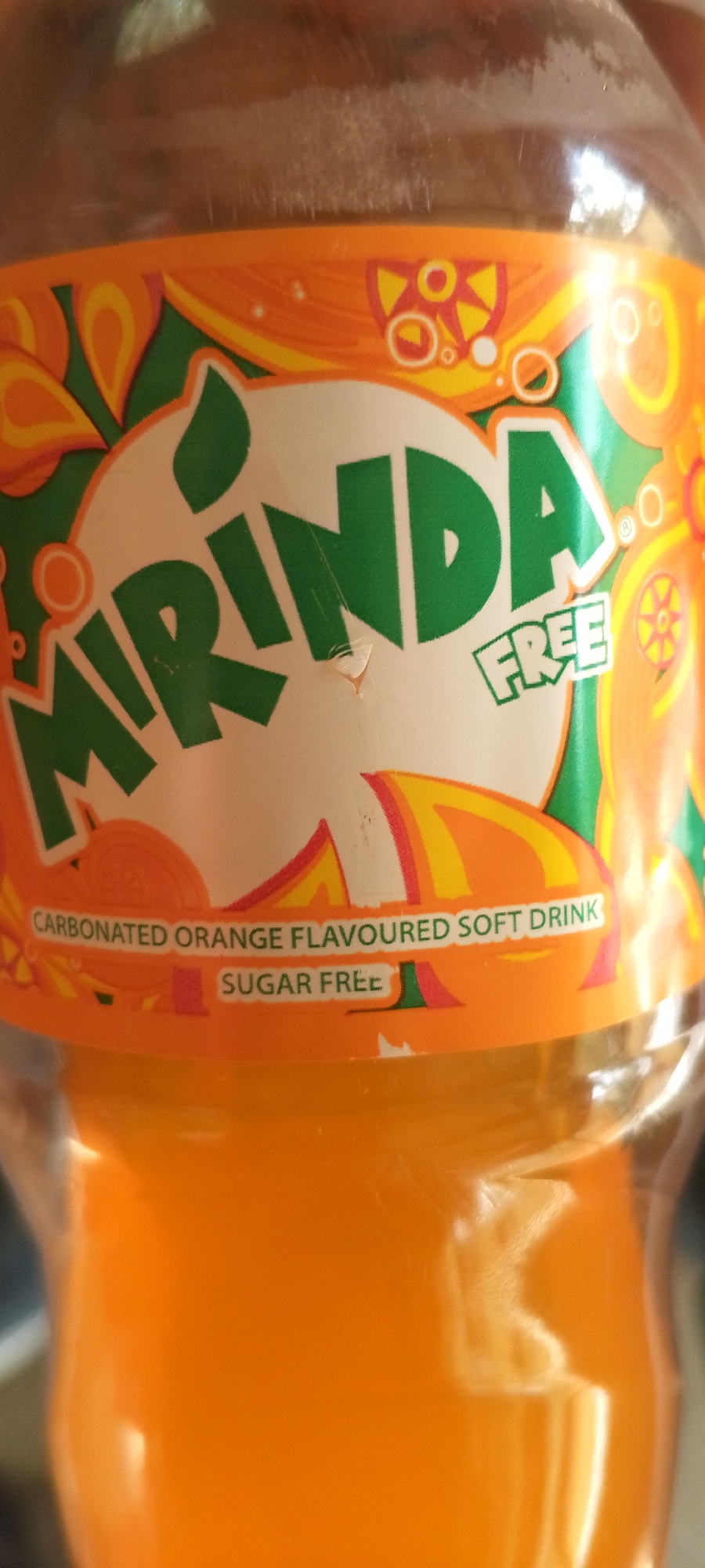 The media file shows a close-up of a plastic bottle of Mirinda, a carbonated orange-flavored soft drink. The bottle has a label with the Mirinda logo in green and white, and the word "FREE" also in green. Below the logo, in smaller white text on a green background, it reads "SUGAR FREE". Further down, on an orange background with decorative swirls, it says "CARBONATED ORANGE FLAVOURED SOFT DRINK". The drink inside the bottle is a vibrant orange color. The lighting suggests it is daytime, and the image appears to be taken indoors or in a shaded area, as there are no harsh shadows. The overall impression is of a product shot, focusing on the branding and attributes of the drink. The location context of Jos, Nigeria, is provided but not visually evident in the image itself.