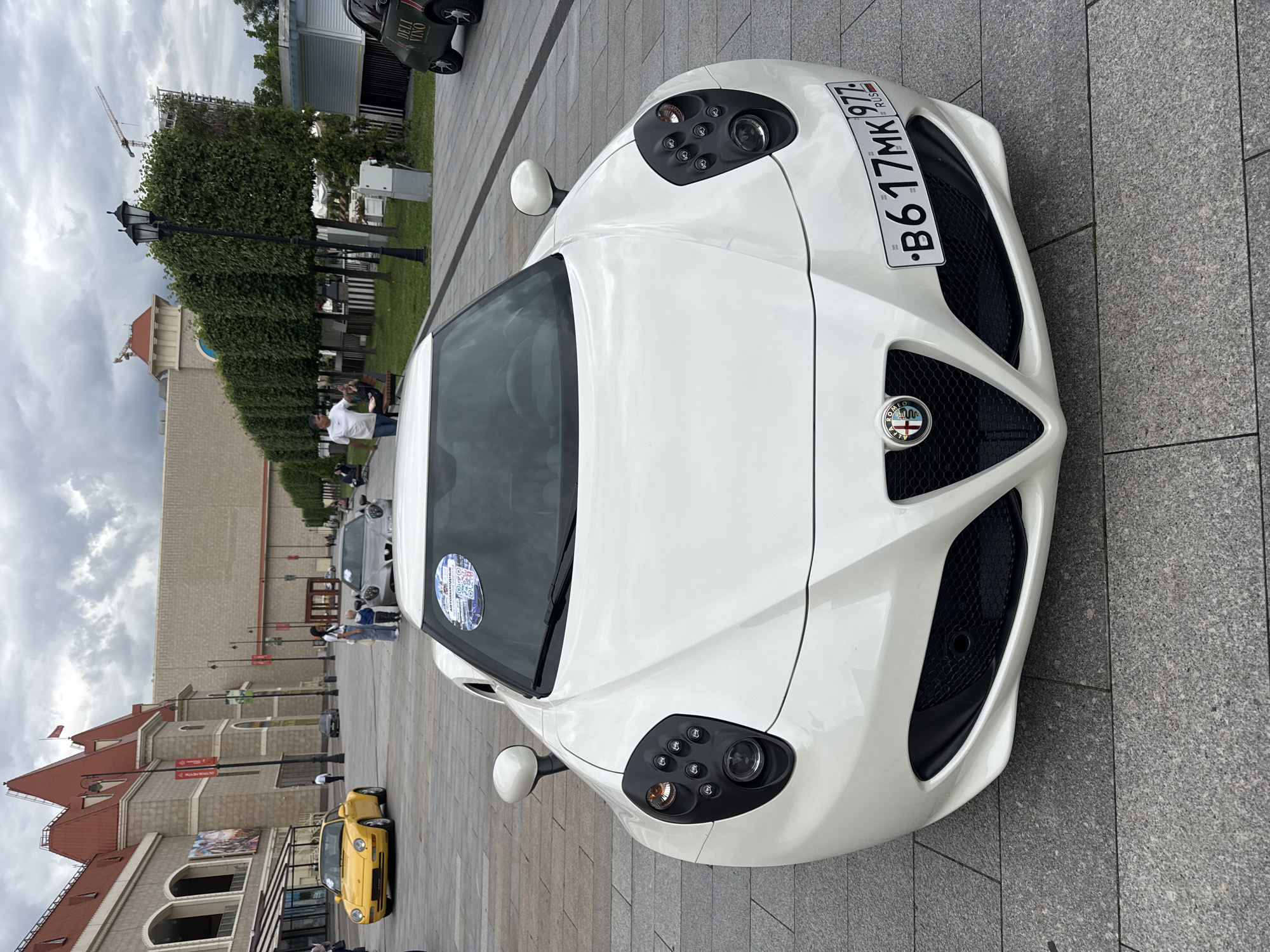 A white Alfa Romeo 4C sports car is parked on a paved outdoor area in Moscow, Russia. The car is clean and shiny, reflecting the overcast sky. A license plate on the front of the car reads "B617MK 977 RUS." A circular sticker with a QR code and some text is affixed to the windshield.
In the background, a yellow Porsche Cayman sports car is visible, as well as other cars and people. A multi-story building with a red-tiled roof and arched windows is to the left of the scene. The atmosphere appears to be casual, with people gathered, possibly for a car show or a similar event. The weather is cloudy, suggesting it might be a mild day. The scene is well-lit, indicating it is daytime.