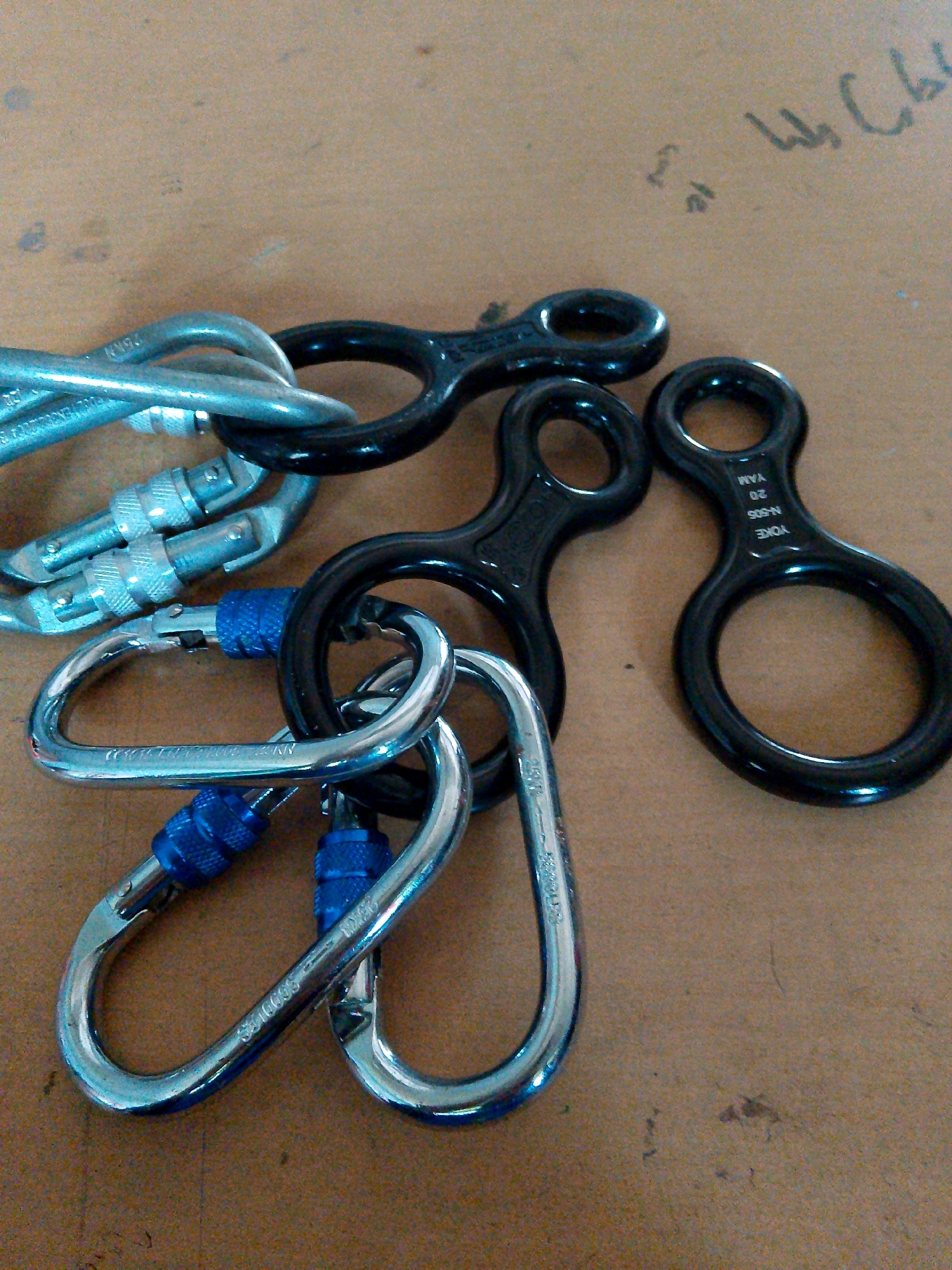 The image displays a close-up, overhead view of several climbing devices, specifically carabiners and "figure-eight" descenders, arranged on a textured, brown surface, possibly a wooden table or countertop. The objects are predominantly metallic, with some featuring blue accents on the carabiner gates.
In the foreground, three silver carabiners with blue locking mechanisms are visible. The carabiners are of varying sizes and shapes, with one larger, D-shaped carabiner positioned prominently in the lower left. Another similar carabiner is placed to its right, and a smaller one is nestled between them. The carabiners appear to be connected to other items, though the full extent of these connections is not clear.
Behind the carabiners, a cluster of black "figure-eight" descenders is arranged. At least three such devices are clearly discernible. These devices are characterized by their distinct shape, with a larger loop at the bottom and a smaller loop above, connected by a central shaft. One of the descenders, in the mid-right of the image, has text engraved on it. The visible text, though partially obscured and possibly upside down, appears to read "YAM 20 N-505-N YOKE".
The background is a mottled brown surface with some subtle variations in texture and color. There are a few dark smudges or specks on the surface, and in the upper right quadrant of the image, some faint, handwritten text or markings are visible. These markings are in a script that appears to be Asian, possibly Indonesian or Malay, but they are too indistinct to be deciphered.
There are no people present in the image. The objects are static, and no actions are being depicted. The lighting suggests an indoor setting, possibly with ambient light. The overall impression is one of organized equipment, likely for climbing or rappelling.
Regarding the location in the city of Kuripan Utara, Indonesia, there is no visual information in the image that can confirm or deny this specific location. The scene is generic in terms of its immediate surroundings and does not contain any landmarks or distinct features that would tie it to Kuripan Utara.