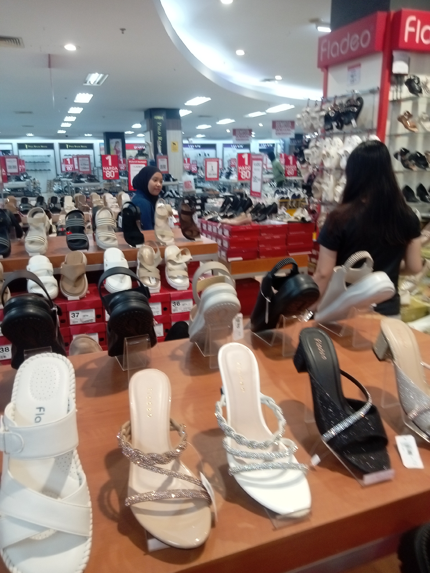 The image shows the interior of a shoe store, likely a Fladeo outlet, in Batam, Indonesia. Several racks and shelves are filled with various styles of women's shoes, including sandals, heels, and flats. Some shoes are displayed on stands, while others are arranged on shelves or in boxes. A woman wearing a blue hijab and a blue jacket is browsing the shoes, while another woman with long dark hair and a dark shirt is visible in the background, also appearing to be shopping. The store appears well-lit with fluorescent ceiling lights. The branding "Fladeo" is visible on signs and some of the shoe displays. The overall environment is that of a retail footwear store.