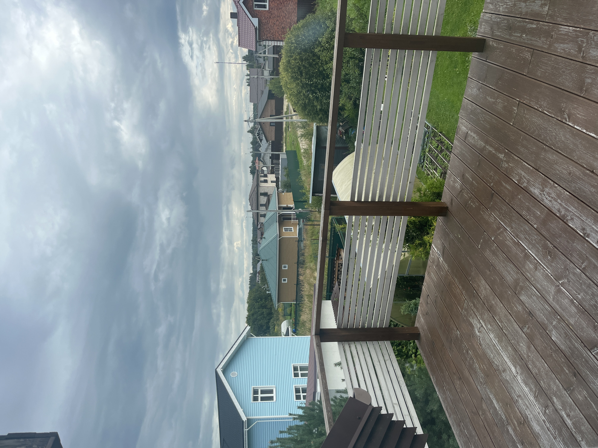 The image depicts a view from a wooden deck overlooking a suburban residential area. The foreground features the brown wooden planks of the deck, with a white slatted railing in the middle ground. Beyond the railing, several houses are visible, including a prominent light blue two-story house on the left and a green-roofed house with a brown facade to its right. Further in the distance, a variety of other houses, trees, and utility poles are scattered across the landscape under a cloudy sky. The overall environment suggests a rural or semi-rural setting in Vladimirovka, Russia, characterized by single-family homes and natural vegetation. There are no people or animals visible in the image.