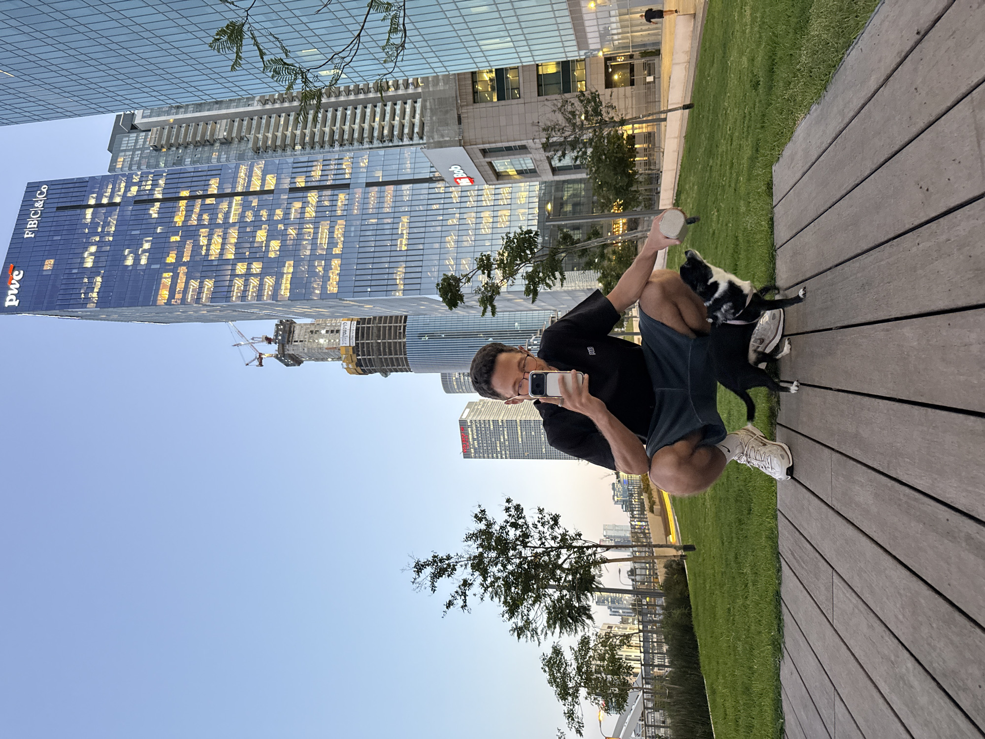 A person squats on a wooden platform next to a grassy area. The person holds a phone in one hand, pointed towards their own face, and a small, cylindrical object in the other hand. A black and white dog stands close to the person. In the background are modern skyscrapers, suggesting an urban setting. Notably, the "PwC" and "FIBI & Co" logos are visible on one building, identifying the location as likely being within or near the Azrieli Center complex in Tel Aviv, Israel. A construction crane is present near one of the buildings. The sky is light blue, indicating either morning or late afternoon.