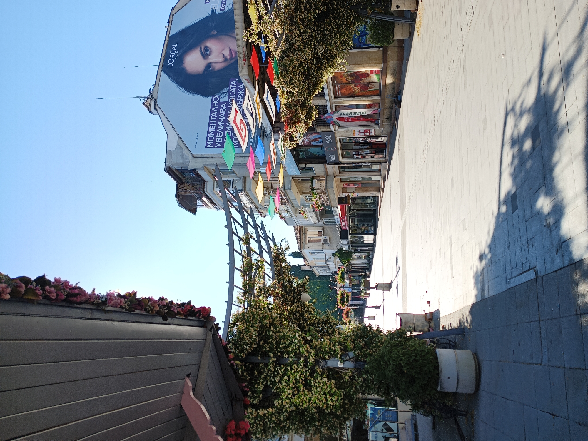 The image shows a sunny day on a pedestrian street in Burgas, Bulgaria. The street is lined with shops and buildings, some adorned with flowers and greenery. Colorful fabric shades are strung across the street, providing shade. A large billboard featuring a woman and L'Oréal Paris branding is visible on a building in the background. The pavement is made of large stone tiles, and shadows of trees and structures are cast across it. There are no people clearly visible in the image. The scene appears to be part of a commercial or tourist area.
