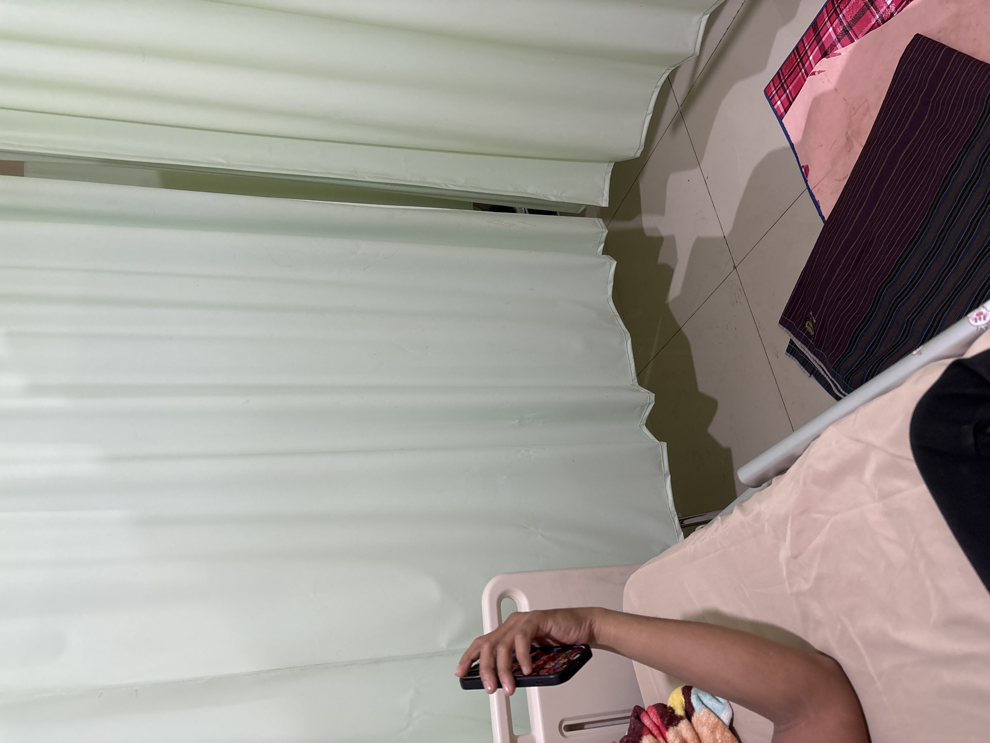A person's arm and hand are visible, holding a cell phone, in a room that appears to be a hospital ward. The person is lying on a bed with light-colored sheets. In the background, pale green curtains are drawn, creating partitions between spaces. The floor is tiled with a light-colored pattern. Two folded fabrics, one dark with stripes and another pink with a plaid pattern, are on the floor. The scene is dimly lit, with shadows cast by the curtains. The location is a hospital ward in Cirebon, Indonesia.