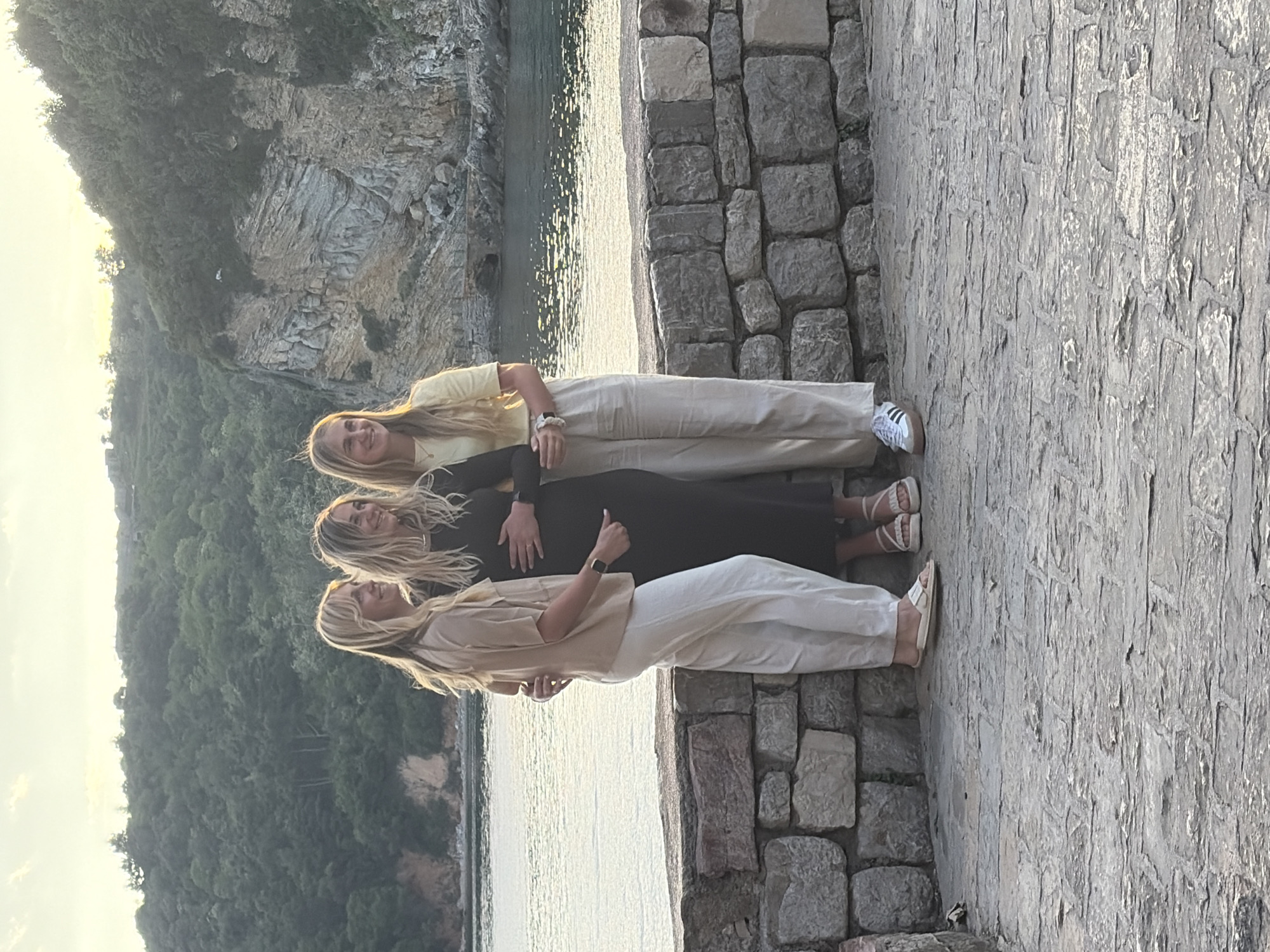 Three women are standing side by side, embracing, on a stone structure with a background of the Adriatic Sea and coastal mountains. The uppermost woman wears a yellow t-shirt and beige pants with white sneakers. The middle woman is wearing a long black dress. The bottom woman wears a beige shirt and white pants. The stone structure they stand on appears to be a protective wall or pier, constructed from roughly hewn stones. Beyond the wall, a body of water stretches towards a landscape featuring greenery and rocky cliffs. In the background, a sunlit sky is partially obscured by the mountains. The location is near the Mogren Fortress in Budva, Montenegro.