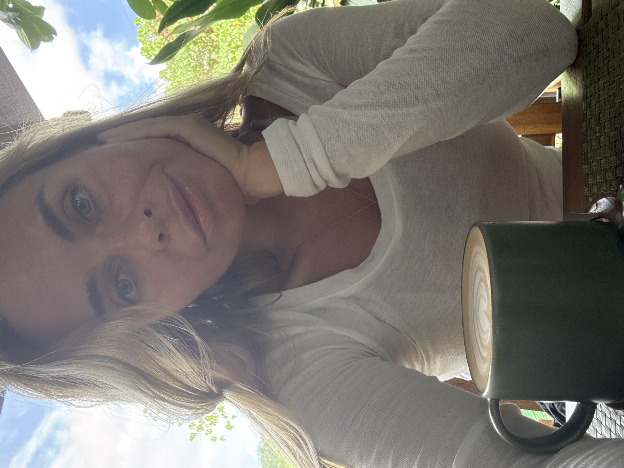 A medium shot photo shows a woman from the chest up, sitting at a table outdoors. She has blonde hair and is wearing a white long-sleeved shirt. Her left hand is resting on her chin, and her right arm is bent at the elbow. In front of her, on the table, is a dark green mug filled with a latte that has latte art on top. The background shows lush green foliage and a blue sky with some clouds, suggesting a tropical outdoor setting. The lighting indicates it is daytime, possibly morning or afternoon. The location is identified as Baan Chaweng Noi, Thailand.