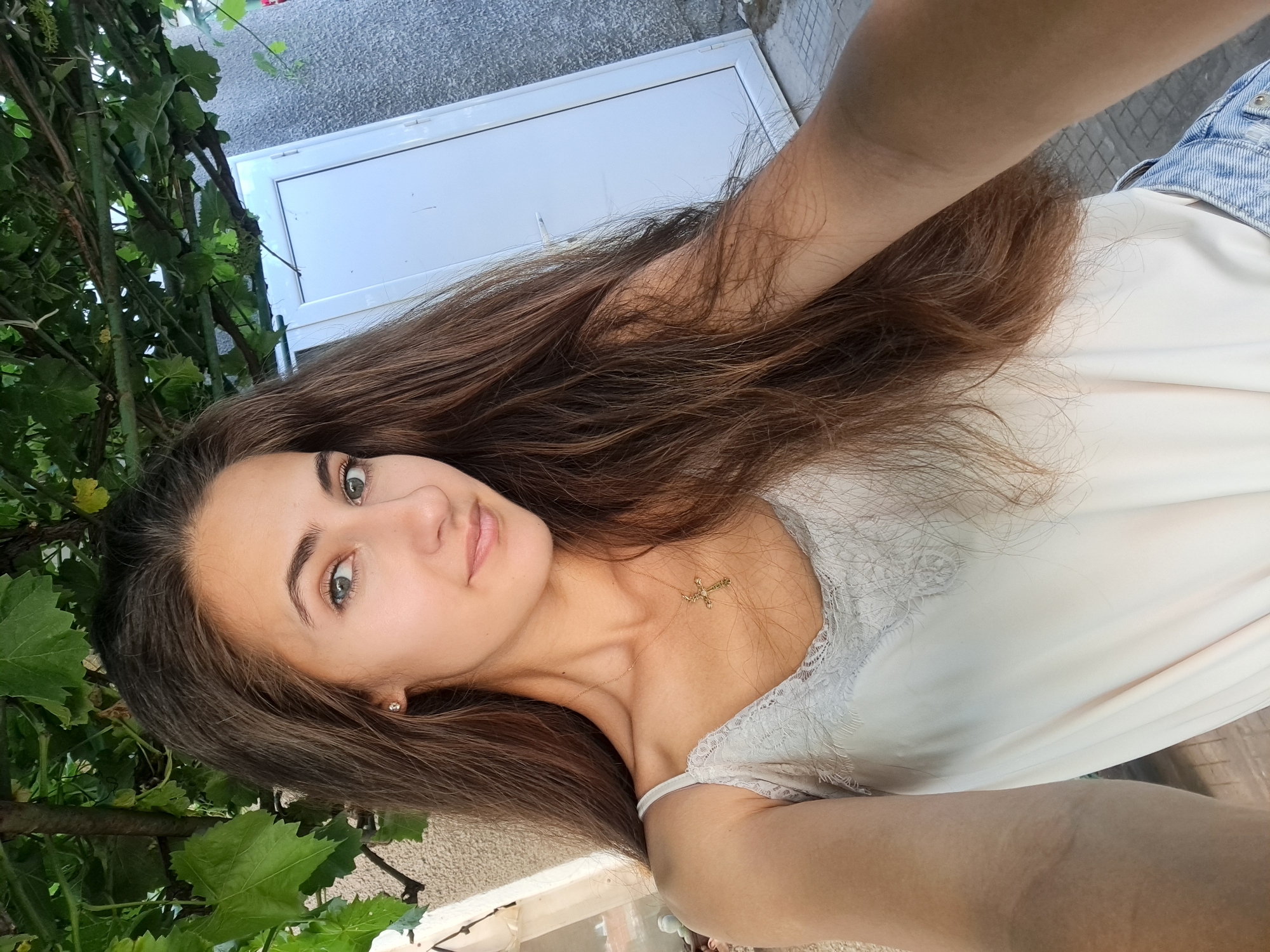 A young woman with long, brown hair is taking a selfie outdoors. She is wearing a light-colored tank top with lace trim and a gold cross necklace. Her hair is falling over her shoulders and partially obscuring her body. Behind her, there is a white door and a textured gray wall, with greenery visible above. The ground is paved with square tiles. The location is identified as Chepintsi, Bulgaria.