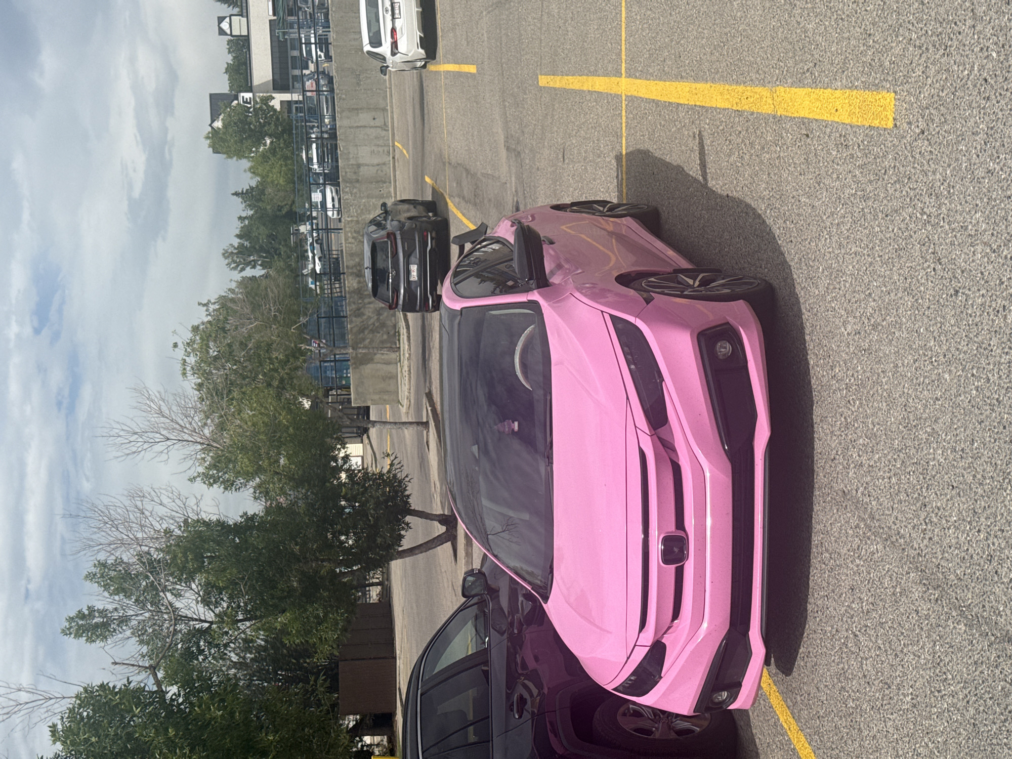 The image shows a bright pink Honda Civic parked in a parking lot. The car has a black roof and black accents on the body, including a spoiler. A dark-colored SUV is parked next to the pink car. In the background, there are trees, a building with a sign that says "E", and other cars parked in the lot. The parking lot is paved and has yellow lines marking the parking spaces. The sky is cloudy. The scene appears to be a typical urban or suburban setting. This image was captured in Calgary, Canada.