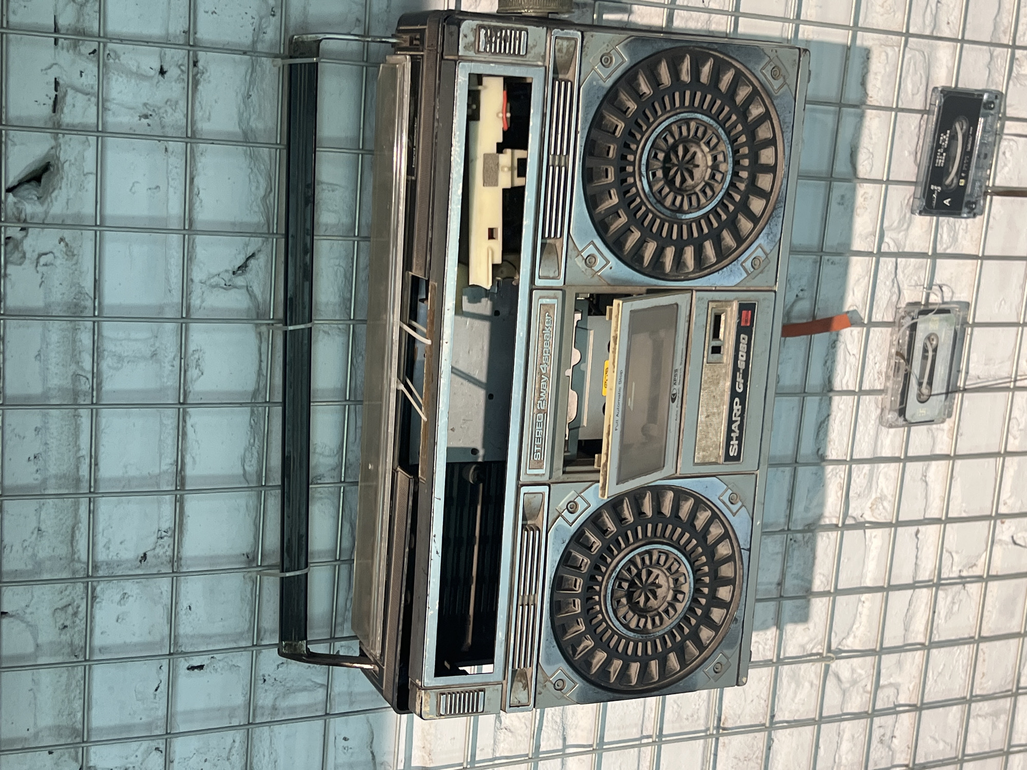 An aged, blue and silver boombox, likely a Sharp GF-6060 model, is mounted on a white brick wall behind a wire grid. The boombox shows signs of wear, with a partially open cassette deck revealing internal components. The lid of the cassette deck is missing. A silver handle is attached to the top of the boombox. Two cassette tapes are displayed below the boombox, also attached to the wire grid. One tape is partially visible on the left, and another, with Chinese text on its label, is visible on the right. The brick wall is textured and appears to be painted a light, somewhat faded color. The wire grid is composed of uniform squares. The lighting suggests an indoor environment, possibly a museum or a retail display. The location is Yuzhong District, China.