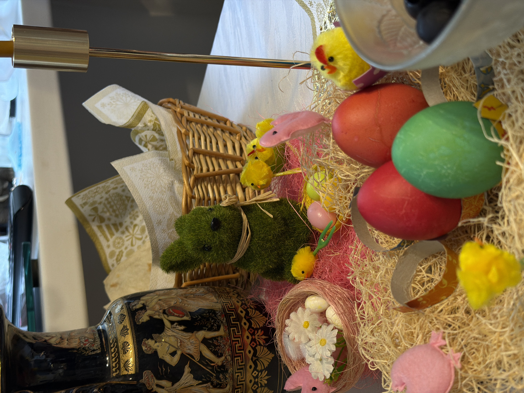 The image is a close-up, slightly angled view of a tabletop Easter display. There are several Easter eggs, some colored red and green, along with yellow chicks, and pink bunny decorations. The eggs and decorations are nestled in straw. A small wicker basket is present, with napkins tucked inside. The base of a black vase with ancient Greek-style figures is visible. The background includes a white tablecloth. There is no evidence in the photo to place this image within Krasnoyarsk, Russia.