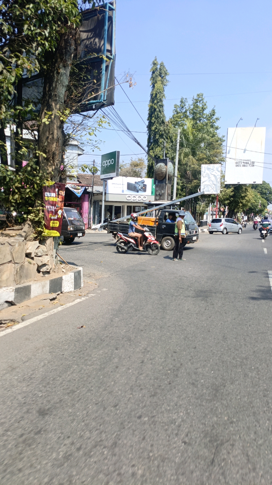 The image depicts a street scene in Banaran, Indonesia, on a clear, sunny day. In the foreground, a paved road curves to the left, with a white and black striped curb on the left edge. A large tree with dense foliage is on the left side of the frame, partially obscuring a billboard structure. To the right of the tree, a vertical banner with the words "GRAND OPENING" and "TERRA COFFEE" is visible.
Further down the road, a black pickup truck with an open bed is stopped. A person wearing a blue shirt and shorts is riding a red and white motorcycle in front of the truck, appearing to be in motion. Another person, wearing a yellow vest and dark pants, stands to the right of the pickup truck, facing the motorcycle. Behind them, a silver car is visible on the right side of the road, and several other motorcycles are further in the distance.
Buildings line the street in the background. One building features prominent signage for "OPPO," an electronics brand, with a green logo and images of smartphones. Other businesses with storefronts are also visible. Overhead, electrical wires and poles crisscross the sky, and several billboards are mounted on structures. Tall, slender trees are interspersed with the buildings, and the sky is a clear blue. The overall impression is of a busy commercial street with ongoing traffic.