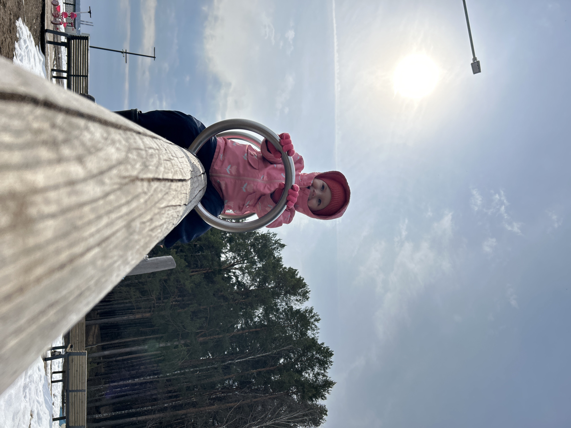 A child in a pink hooded jacket and gloves plays on a seesaw at a playground in Chaikovsky, Russia. The seesaw is made of wood and metal. A bench and trees are visible in the background. The sky is cloudy with the sun shining through.