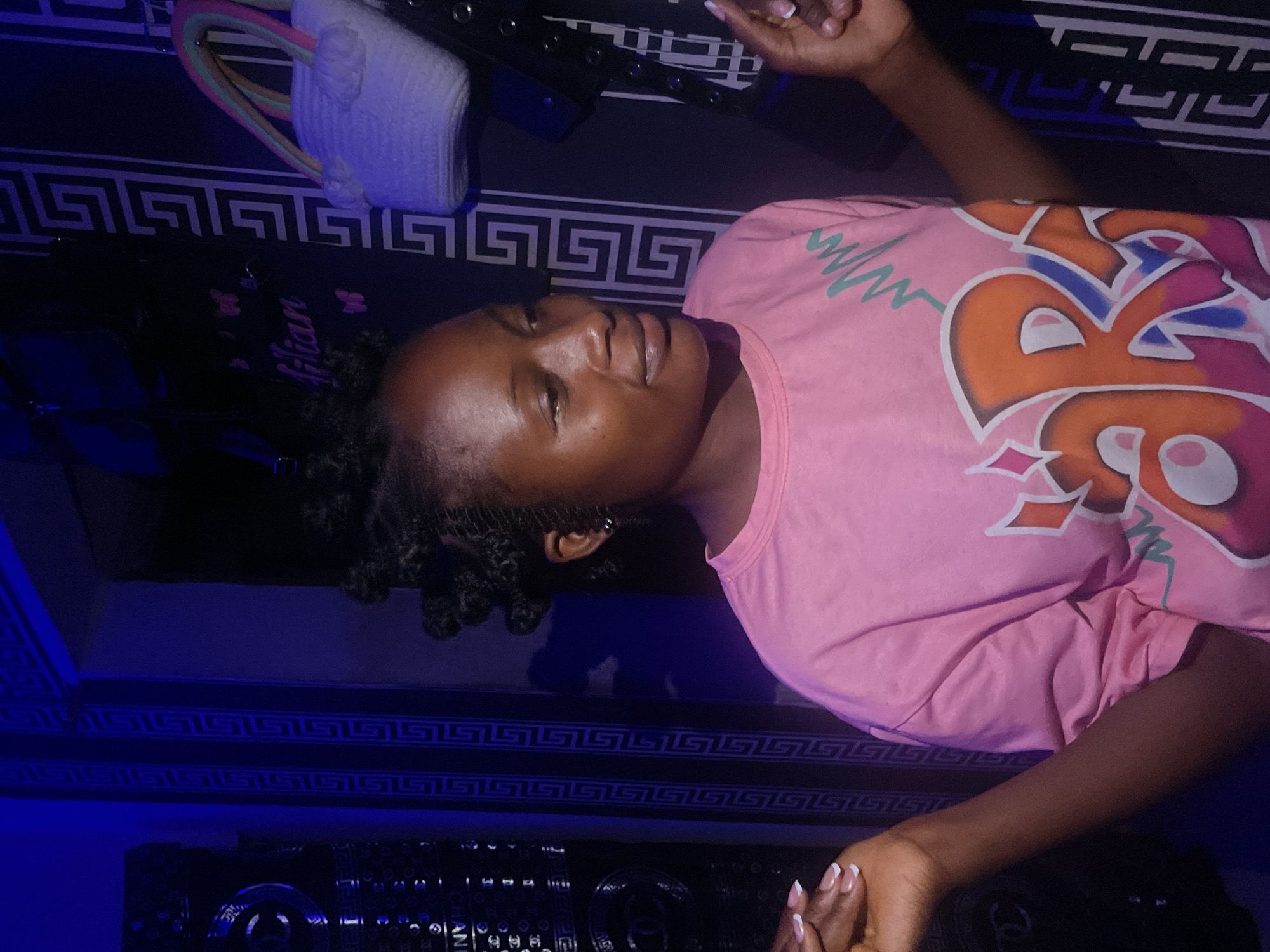 A young woman is lying down in an indoor space. She is wearing a pink graphic t-shirt. Her hair is styled in small braids or twists. Her fingernails are painted with a French manicure. The wall behind her features a black-and-white Greek key pattern. There are clothes hangers and clothing visible in the upper portion of the image. The lighting appears to be dim, possibly with a blue hue. The location is likely a room in Lokoja, Nigeria, based on the prompt, but there is no further information visible in the image to specifically confirm this.