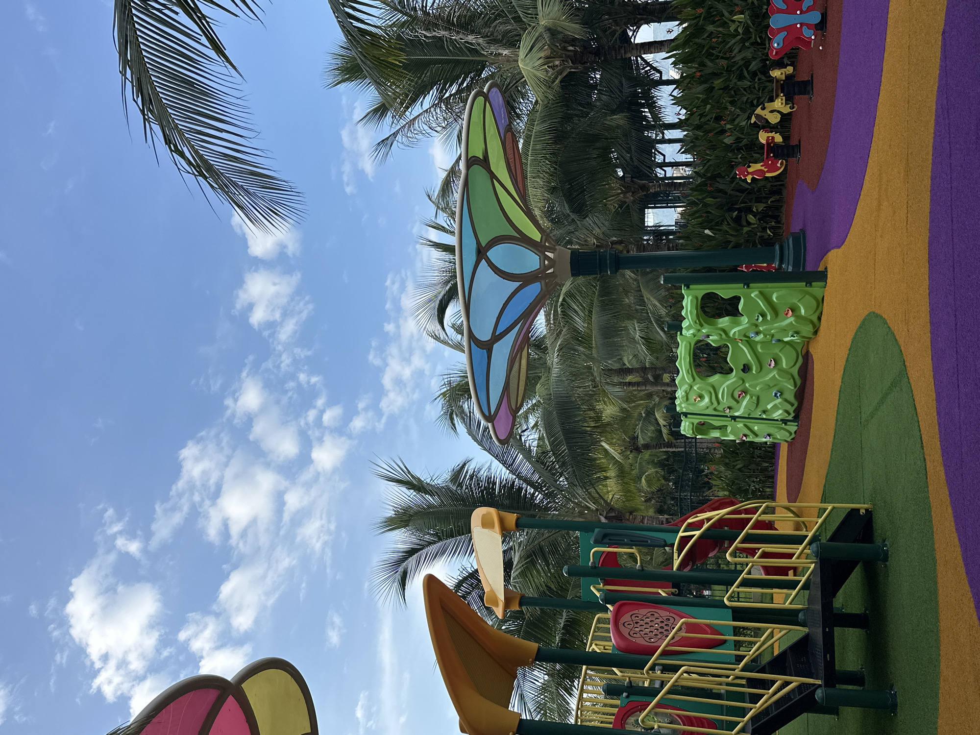 This image depicts an outdoor playground located in Nha Trang Ward, Vietnam, under a bright blue sky with scattered white cumulus clouds.
In the foreground on the left, a multi-level playground structure is visible, featuring dark green support posts, bright yellow metal railings, and a black staircase. Red and green panels are integrated into the structure, and yellow canopy-like elements are present at the top. The ground immediately surrounding this structure is covered in dark green artificial turf.
To the right, a freestanding green climbing wall with various colorful handholds is situated on a reddish-brown soft-fall surface. Further right, three spring riders are positioned: one shaped like a red rooster, one like a yellow horse, and one like a red and blue butterfly, all resting on the reddish-brown surface.
The ground throughout the playground consists of a varied pattern of soft-fall materials, including sections of dark green artificial turf and curved areas of yellow, purple, and reddish-brown textured surfacing.
A prominent, decorative, umbrella-like structure, possibly a shade or lighting feature, stands near the center-right of the image. It features a multi-colored top with segments resembling stained glass in shades of blue, green, purple, and pink, supported by a dark green pole.
The background is dominated by lush tropical vegetation, including numerous tall palm trees with green fronds and dense bushes with dark green leaves, some exhibiting small red flowers. A dark fence is partially visible through the foliage in the background. Hints of a body of water or distant land features can be discerned between the trees on the horizon. No individuals are present in the scene. The lighting indicates clear daylight conditions.