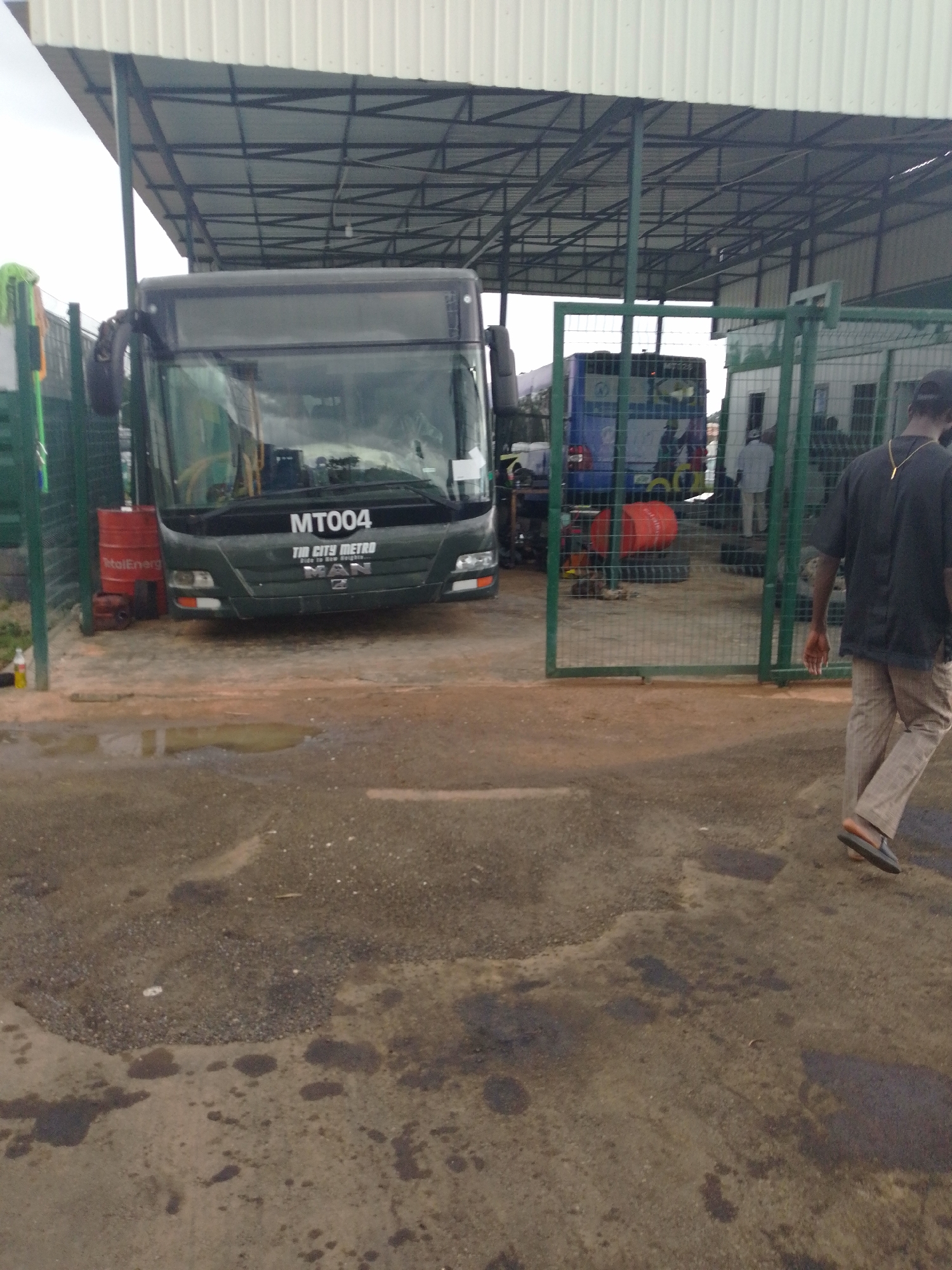 An exterior, medium-angle, eye-level shot captures a scene under a covered structure, likely a bus depot or maintenance area, in Barikin Jirga Summa, Nigeria. In the foreground, a man walks away from the camera, his back to the viewer. He wears a dark short-sleeved shirt, tan pants, and dark sandals. His right hand is extended slightly, palm facing forward. The ground is paved with a mixture of asphalt and gravel, with several dark, oily patches and a few puddles reflecting the overcast sky.
Dominating the mid-ground is a dark green bus with the identification "MT004" and "THE CITY METRO" displayed on its front. The bus appears to be parked, its headlights visible. To the left of the bus, a bright red barrel with "TotalEnerg" printed on it stands on the ground. Beyond the bus, through a green metal fence, another blue bus is partially visible, along with a small white building. Several individuals, appearing to be men in light-colored clothing, are seen near the building and the blue bus, suggesting they are staff or workers. The overall environment appears functional and utilitarian, with the presence of buses, maintenance equipment, and personnel.
