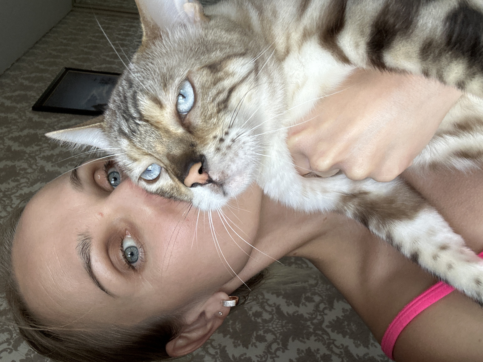 A woman with light brown hair and blue eyes is holding a cat with blue eyes and brown markings. The woman is wearing a pink top. The cat is being held close to the woman's face, and it appears she is kissing the cat. The background is a wall with a floral pattern. The image appears to be taken indoors. There is a framed picture on the wall in the background. No specific location in Energetik, Russia is discernible.