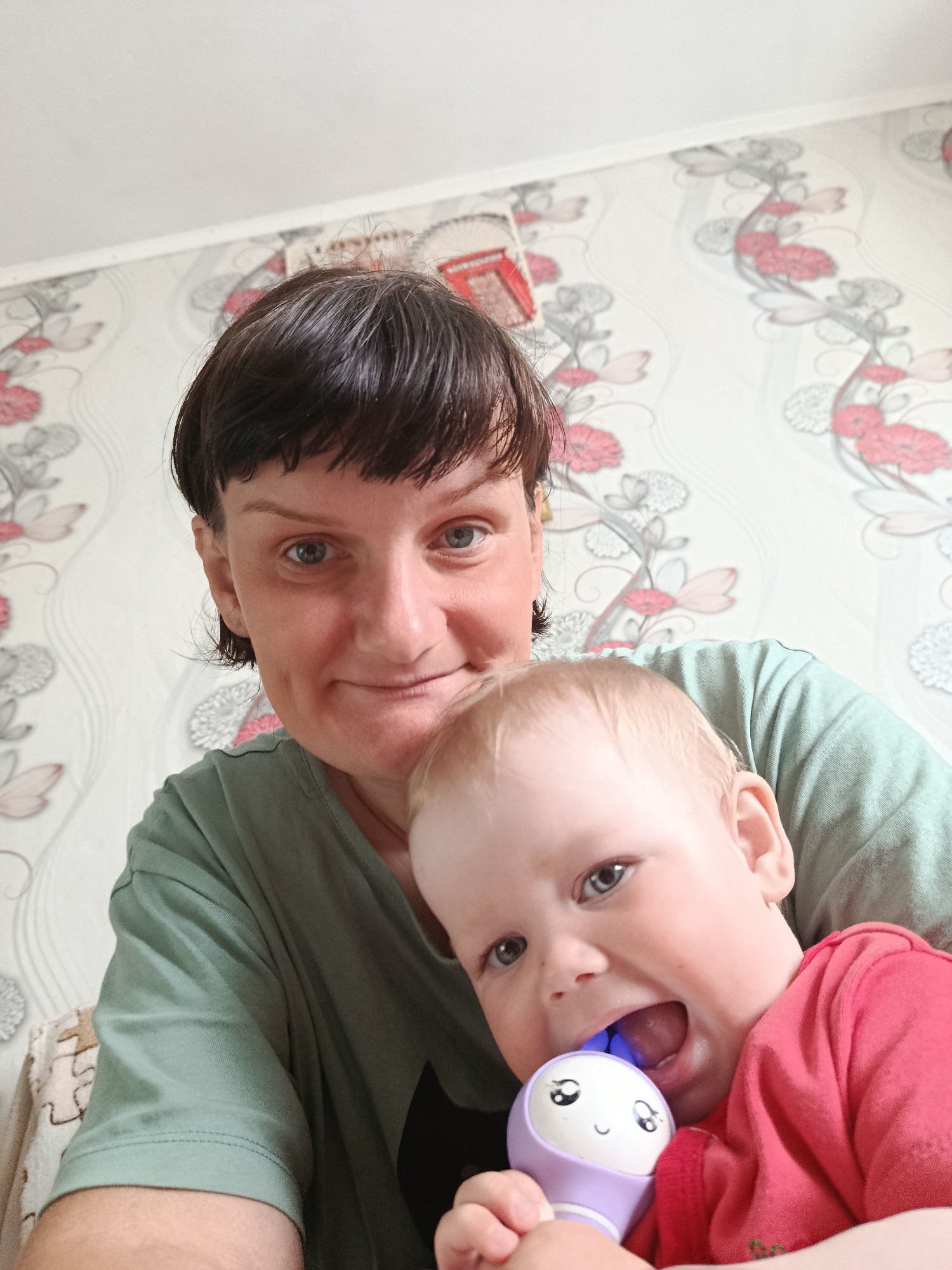 This close-up indoor image features an adult woman and a baby, likely a mother and child, captured in what appears to be a selfie. The scene is set inside a home in Foky, Russia, against a wall covered with wallpaper featuring a repeating pattern of stylized red and pink flowers with grey-white foliage on a light background.
The woman, positioned behind the child, has short dark hair with bangs and light blue eyes. She wears a plain green t-shirt and has a slight, calm smile or smirk on her face as she looks towards the camera.
The baby, appearing to be a toddler, is in front of the woman and is actively engaged with a toy. The child has light blonde hair and light blue eyes, and wears a red t-shirt. Their mouth is open wide, revealing a blue light emanating from inside a purple and white toy held up to their mouth. The toy has a cheerful, cartoon-like smiling face on its white upper portion. The child also looks towards the camera, seemingly playful or curious.
The lighting in the image is bright and even, suggesting it is daytime. The moment appears informal and warm, capturing an everyday interaction between the two individuals. No visible text is present in the image.