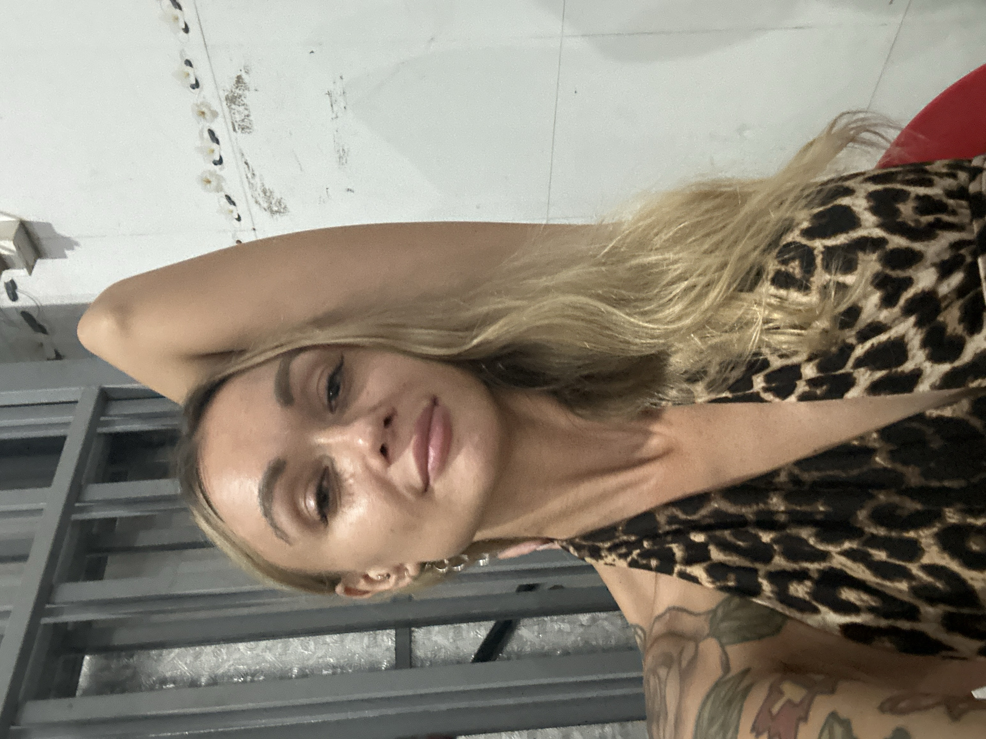 A woman with blonde hair and a leopard-print top is shown in a close-up shot. She has tattoos on her left arm and is wearing jewelry in her ears. She is indoors, standing near a metal structure with what appears to be a white wall in the background. The photo was taken in Phu Thuy Ward, Vietnam.
