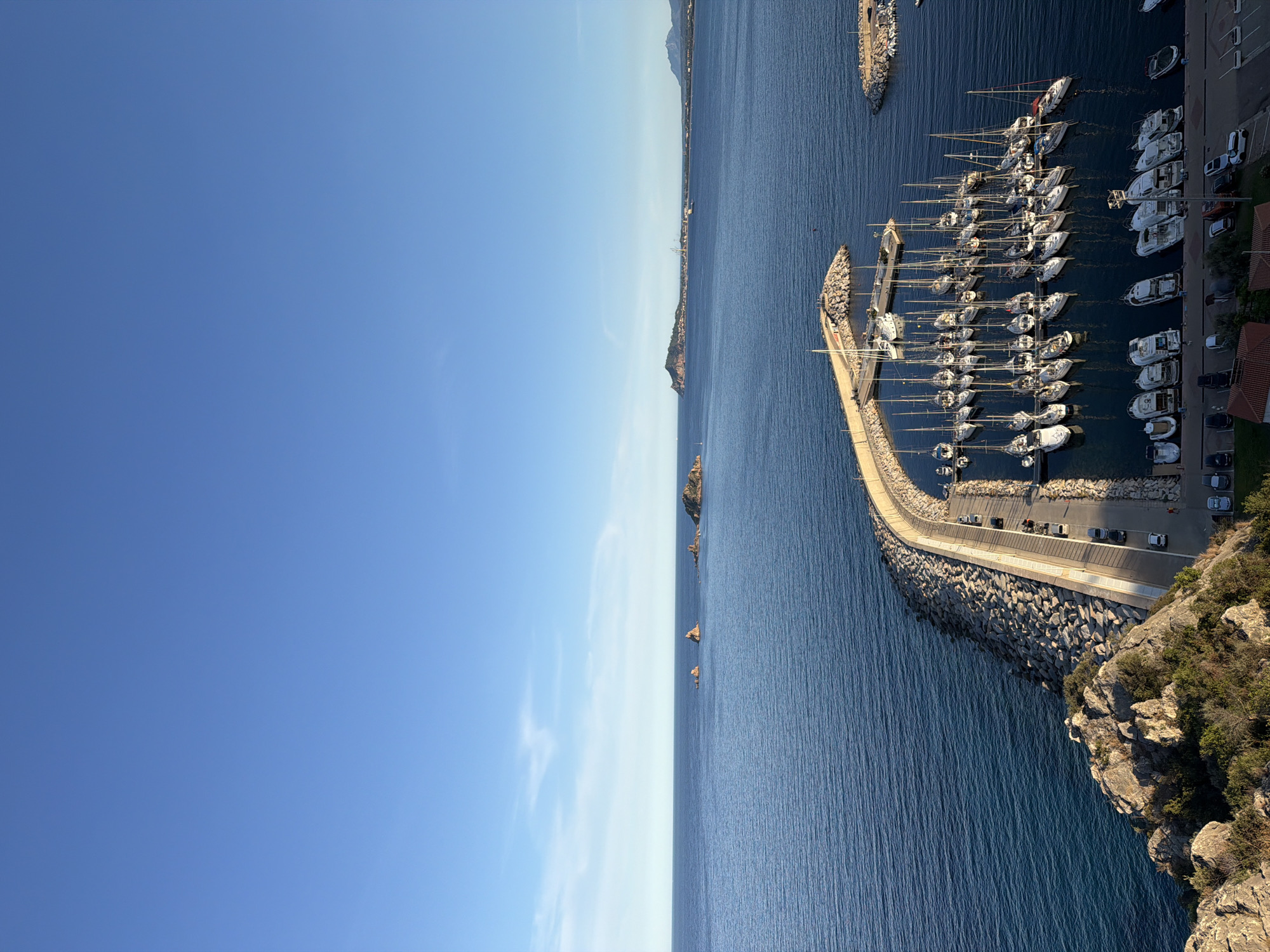 The image shows a high-angle view of a marina in Santa Maria Navarrese, Italy. The marina is filled with numerous sailboats and yachts docked in calm, blue water. A curved concrete and stone breakwater extends into the sea, protecting the harbor. Along the breakwater, cars are parked on a paved road. In the background, a vast expanse of blue sea meets a clear sky, with distant islands and a coastline visible on the horizon. The overall scene depicts a serene coastal landscape with a bustling marina.
