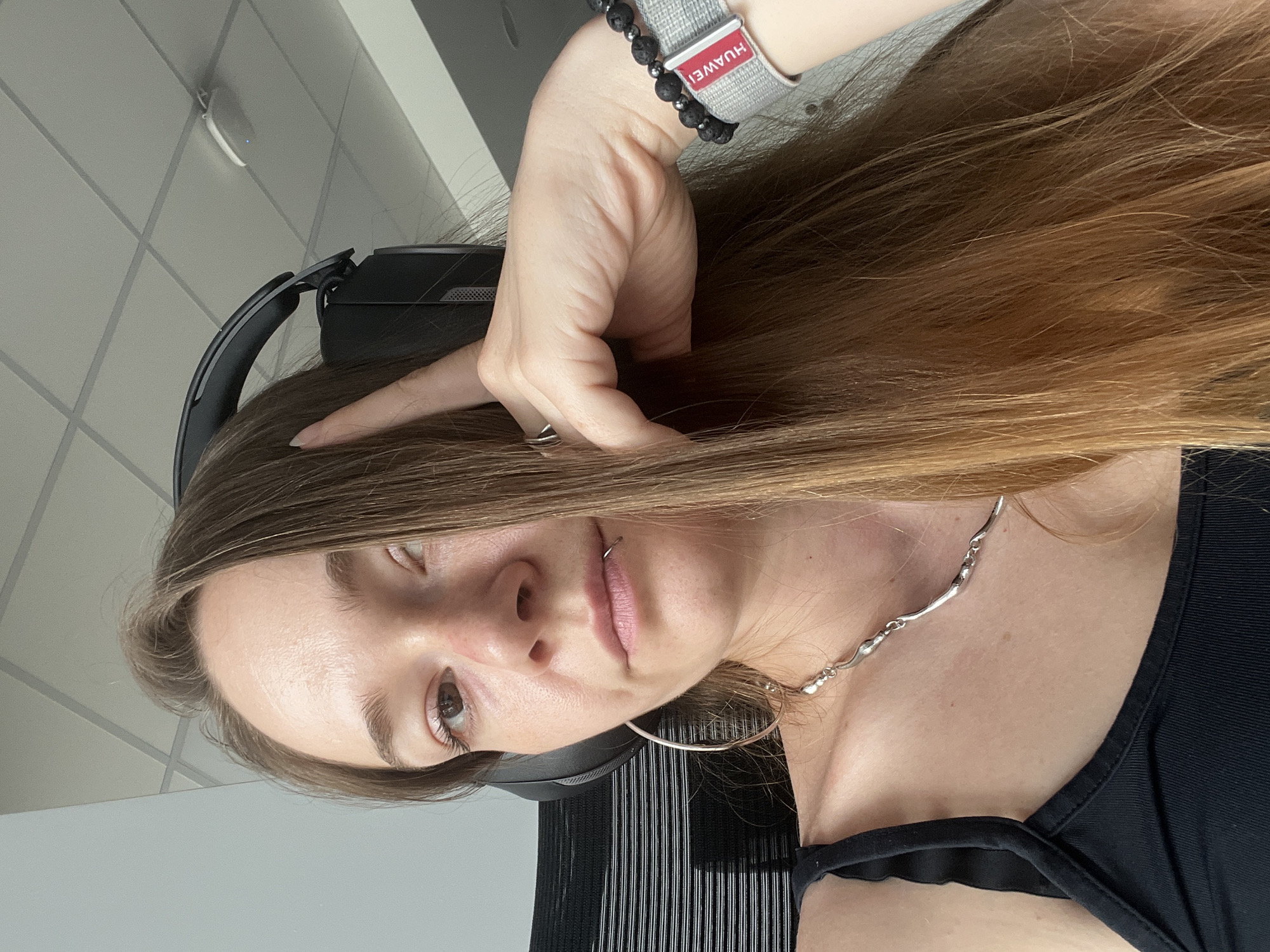 This media file features a young woman in a close-up, selfie-style shot, oriented sideways. She has long, light brown hair that cascades over her shoulder. She is wearing black headphones, a black tank top, and a silver necklace. A notable detail is a bracelet on her left wrist that has a grey fabric band with a red tag that says "HUAWEI". She also has a ring on her left hand and a lip ring. The woman's expression is neutral, with her eyes looking towards the camera.
The setting appears to be indoors, likely an office or a similar environment, suggested by the tiled ceiling and a wall in the background. There's a suggestion of fluorescent lighting. The time of day and weather are not discernible from the image. The overall mood is casual and perhaps contemplative, given her posture and the way she is touching her head with her hand. No specific event is taking place, it appears to be a candid or posed portrait. The location context of Plovdiv, Bulgaria, does not provide any visual cues within the image itself.