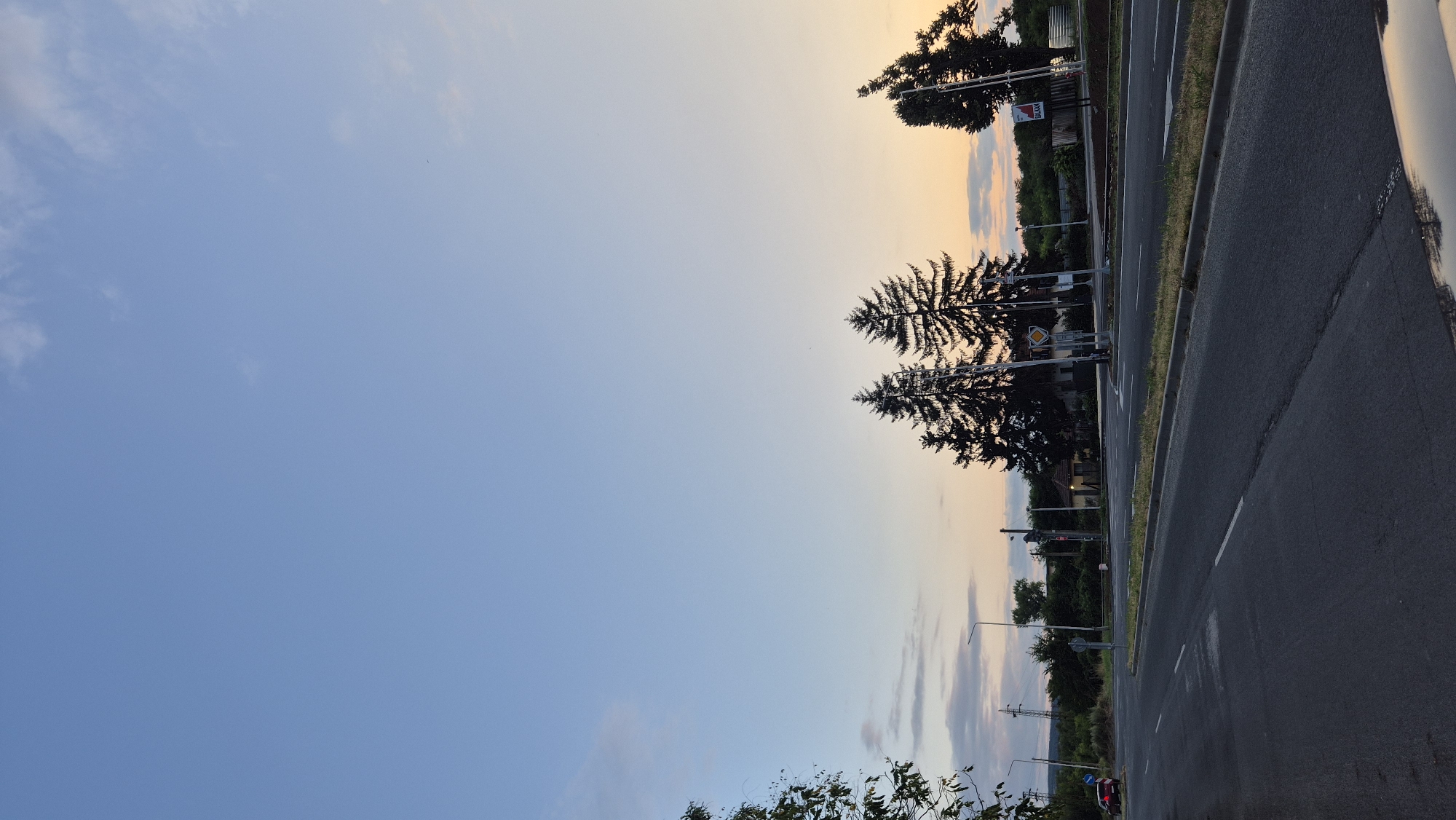 A low-angle, vertical, outdoor, full shot captures a road intersection at dusk in Lovech, Bulgaria. The sky transitions from a light blue at the top to a soft orange-pink hue near the horizon, with faint clouds scattered throughout. In the middle ground, a cluster of tall, dark evergreen trees stands silhouetted against the colorful sky, with a smaller evergreen to the far right. A road with white lane markings curves through the foreground, with a grass median separating it from lanes on the right. Various traffic signals, poles, and a small building are visible behind the median, along with distant vegetation and power lines. A red car is parked on the left side of the intersection, and a sign with the word "BALKAN" in red lettering is visible on the right. The overall atmosphere is serene and transitional, marking the end of the day.