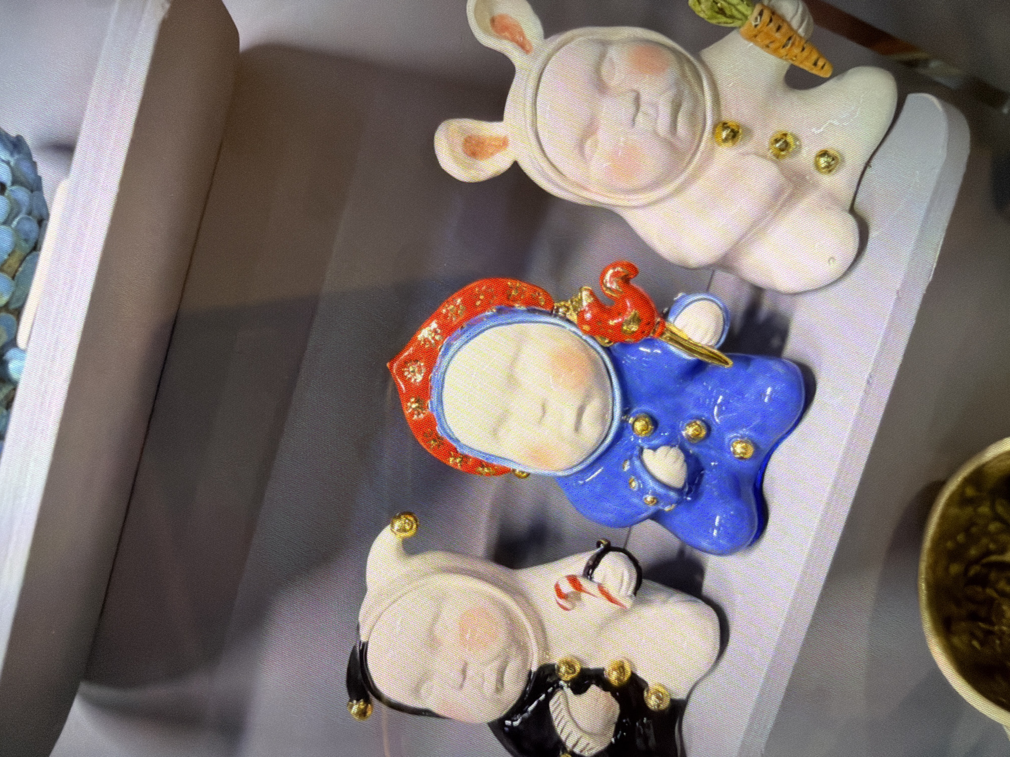 Three decorative ceramic figures are prominently displayed on a light-colored shelf, viewed from a slightly elevated angle.
The uppermost figure is a white rabbit, depicted lying down on its back with its arms raised. It has pink inner ears and rosy cheeks, and holds a small, textured orange carrot in one hand. Gold, bead-like embellishments are scattered across its body. Its facial expression appears content and peaceful.
Below the rabbit, positioned centrally, is a blue figure with a distinctly rounded, somewhat star-like or cloud-like shape. This figure is adorned with a red hood featuring gold details and white trim, and its face shows closed eyes, suggesting sleep or serenity. A small, intricately detailed red bird with gold accents rests near its head. More gold, bead-like details are visible on its blue body.
The lowermost figure is also white, with black and white striped accents on its ears and a black lower body section. A small red and white striped object, resembling a candy cane, is placed near its head. This figure also features multiple gold, bead-like adornments and has a calm, smiling expression.
All figures appear to be glazed, giving them a smooth, glossy finish. They are static objects, arranged on a pale grey display surface, possibly a shelf or cabinet. On the far left, a portion of another textured, possibly blue-green object is visible. On the far right, part of a brownish-green, bowl-like object can be seen. The background is a plain, light grey wall. There is no visible text in the image. The overall impression is one of charming, handcrafted decorative items.