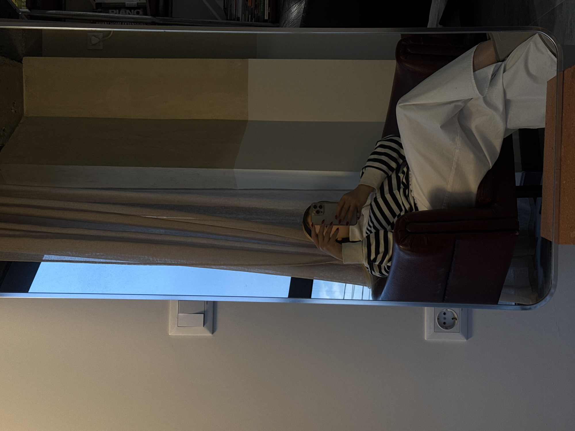The image is a reflection in a mirror. In the mirror's reflection, a person is sitting in a leather chair, wearing white pants and a striped shirt. They are holding a phone in their hands. The background includes a shelf with books, a beige wall, and a curtain. The location appears to be indoors, likely in a living space. Electrical outlets and light switches are visible on the wall where the mirror is hung. There is no definitive information to confirm the location is Yerevan, Armenia.