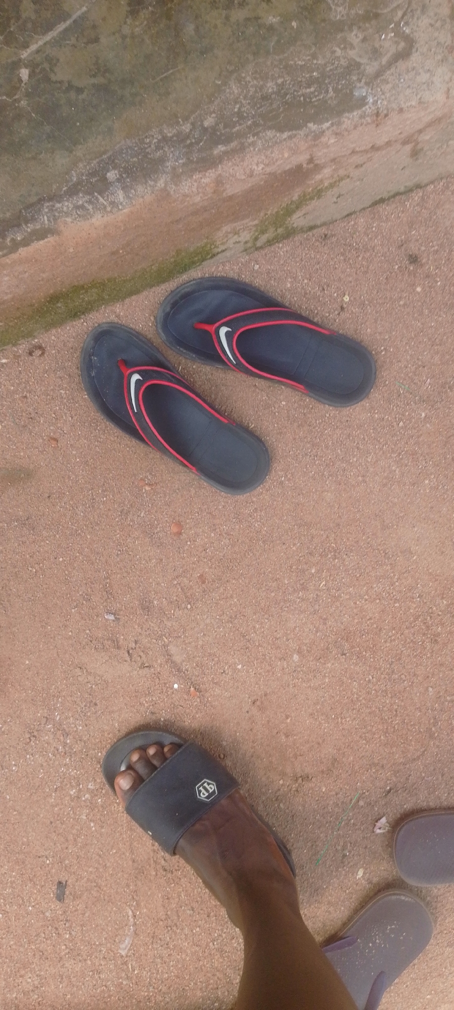 The image shows a person's feet wearing footwear on a sandy ground. In the foreground, a person's right foot is visible, wearing a black slide sandal with a silver emblem. To the left of this, another black slide sandal is partially visible. Further up in the frame, a pair of dark blue flip-flops with red trim and white Nike swoosh logos are lying on the ground. The ground appears to be a mix of sand and dirt, with some small pebbles and debris. In the upper left portion of the image, a concrete structure with a green algae-like growth is visible, suggesting an outdoor setting. The lighting suggests it is daytime. No specific activity or event is clearly depicted, beyond the presence of footwear. The location cues are general to an outdoor, possibly somewhat unkempt, area.