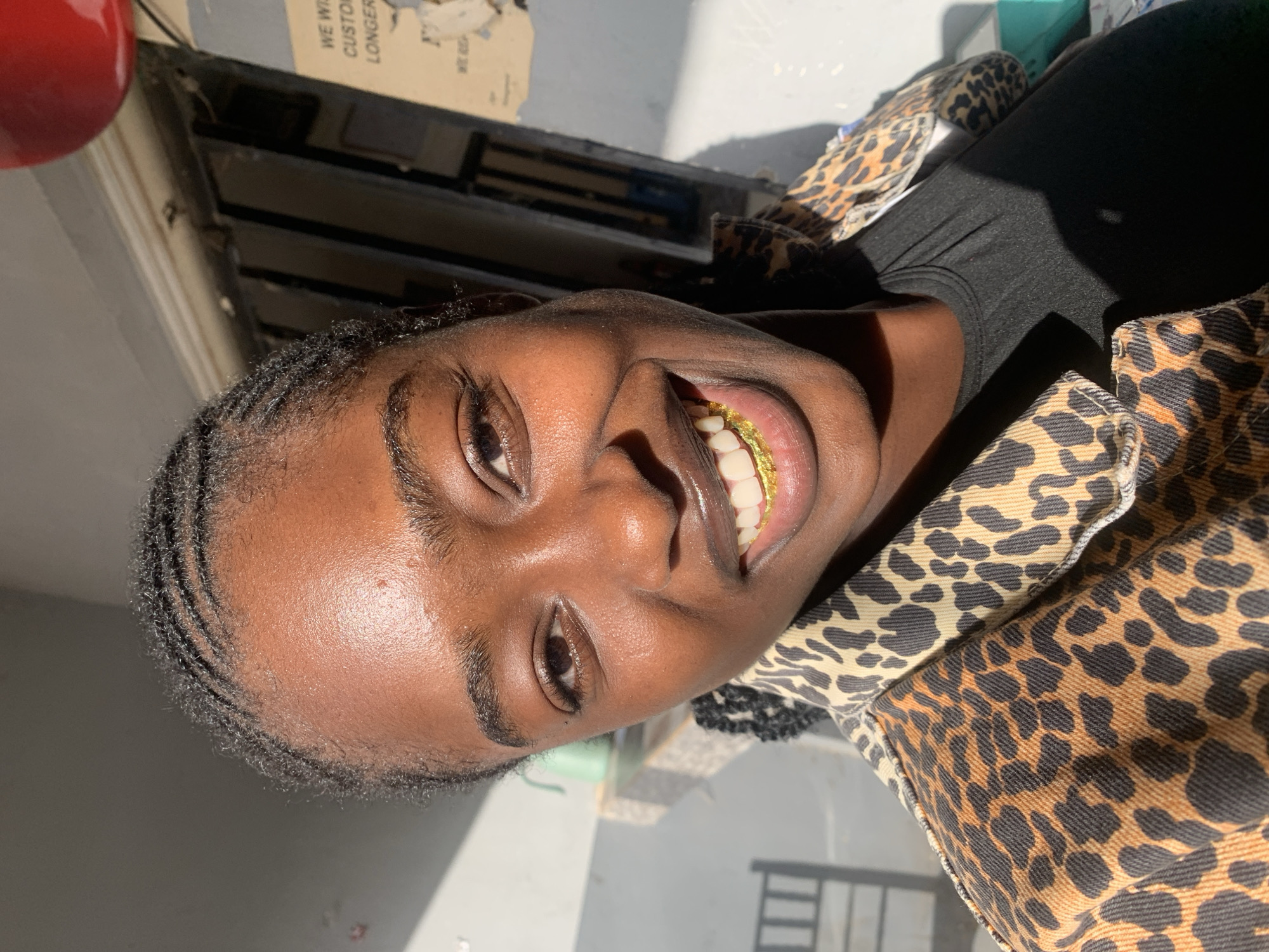 The image, reportedly captured in the city of New Stadium, Nigeria, features a young Black woman smiling broadly directly at the camera. Her dark hair is styled in cornrows, pulled back from her face. She is wearing a light brown and dark brown leopard print denim jacket over a plain black t-shirt or tank top. A prominent gold-colored dental accessory, resembling foil or a partial grill, covers her lower front teeth, visible as she smiles. Strong natural light from the left illuminates her face, creating distinct highlights on her forehead, nose, and cheeks, and casting a pronounced shadow across her right neck and chest.
The background on the left side of the frame includes a light grey wall or ceiling, a light turquoise object (possibly a cup or container) resting on a white ledge, and a partial view of a dark metal chair back. The background on the right reveals a distressed, light-colored wall with peeling paper or paint, revealing darker layers beneath. Fragments of text, including "WE WILL CUSTOM LONGER," are visible on this wall. Further back on the right, dark, rectangular recessed openings (possibly windows or shelves) are present, and a red spherical or bulbous object (such as a balloon or light fixture) is visible in the upper right quadrant of the frame.