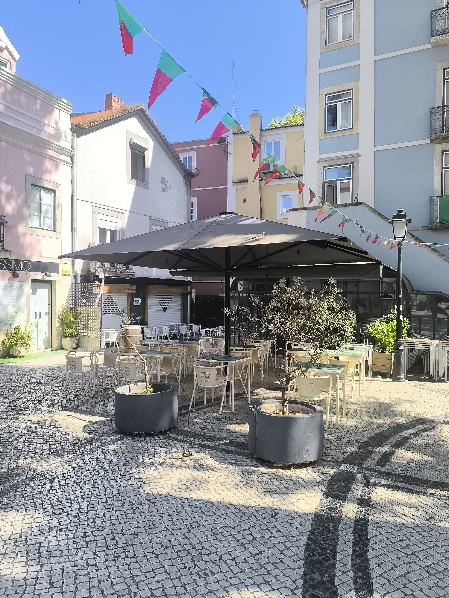 The image depicts an outdoor seating area of a cafe or restaurant located in Setúbal, Portugal, likely in a public square or a pedestrian street. The ground is paved with traditional Portuguese cobblestones, a pattern of black and white stones forming a textured surface.
Dominating the scene is a large, dark grey umbrella, providing shade over a collection of tables and chairs. The tables have light-colored tops, possibly green or white, and are surrounded by white chairs. Some chairs have the imprint of a logo that appears to read "SAGRES", a Portuguese beer brand. In the foreground, a cylindrical grey planter contains a small, leafless tree. Another similar planter is visible to the left, with a small plant.
In the background, several buildings line the square. To the left, a building with a pink facade has a store with a sign reading "VISSIMO". Further back, there are buildings in pastel yellow, pink, and blue colors, all with windows and balconies. Strings of triangular flags in green and red, resembling the Portuguese flag, are strung across the sky between the buildings, suggesting a festive occasion.
A black lamppost stands to the right of the frame, contributing to the street furniture. Several stacks of chairs are visible in the far right, indicating that the area may be closed or prepared for a different setup. Shadows from the umbrella and buildings cast patterns on the cobblestone ground, highlighting the bright, sunny day. The sky is a clear, vibrant blue. There are no people visible in the image, giving the scene a quiet, unoccupied atmosphere.