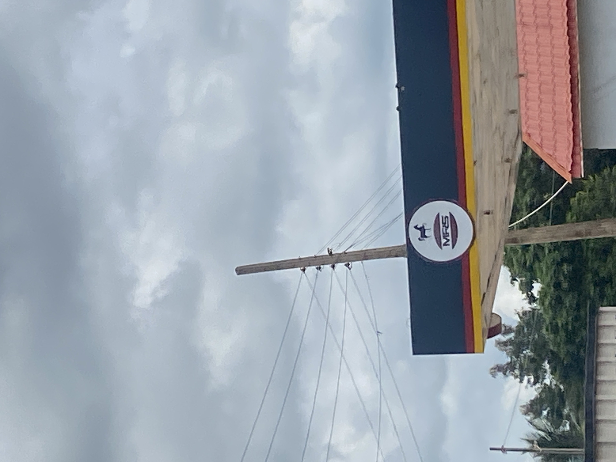 This image, taken in Sasaro, Nigeria, captures an outdoor scene under an overcast sky filled with light grey clouds. The main subject is a portion of a commercial building, likely a gas station canopy, shown horizontally across the middle of the frame (if rotated upright). This structure features a prominent dark blue section with a yellow and a narrower red stripe running horizontally, and a lighter-colored beam or support beneath.
A circular white sign is affixed to the structure, displaying the text "MRS" and a logo resembling a deer or antelope. To the left of this sign, a wooden utility pole extends diagonally, from which several power lines or guy wires descend.
In the upper right background, a section of a red-tiled roof is visible, suggesting an adjacent building. Beneath this, vibrant green foliage from trees fills the background. A partial view of a white wall or structure is also visible in the bottom right corner. The image depicts a static, daytime scene with no discernible activity.