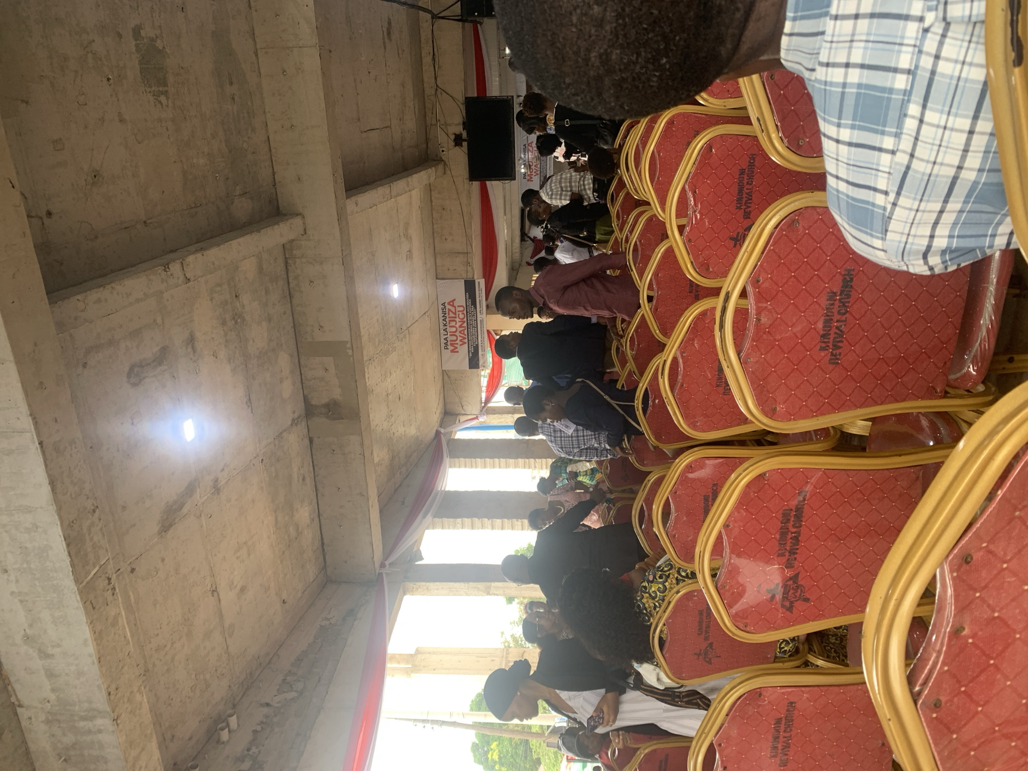 The image shows a gathering inside a large, unfinished-looking building, presumably in Dar es-Salaam, Tanzania, given the signage visible in the scene. The building appears to be a church. Rows of red chairs with gold frames are arranged for attendees. People are seated and standing, engaging in the event. A banner or sign with the words "PAA LA KANISA MUUJIZA WANGU KINONDONI REVIVAL CHURCH" is visible at the front of the room. A screen is also present. The overall impression is a religious gathering in a structure that is still under construction or has an intentionally unfinished aesthetic.