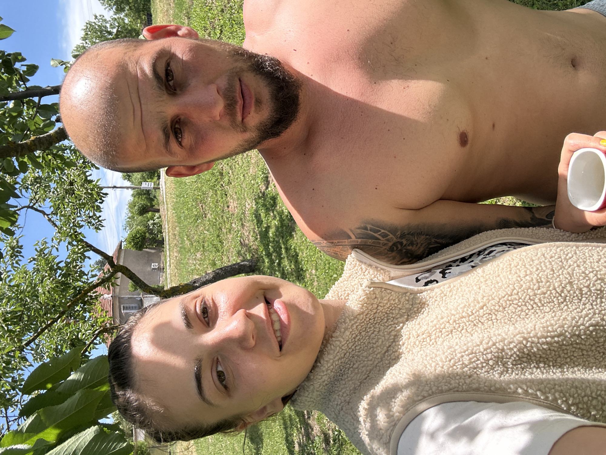 A photograph taken in Sirakovo, Bulgaria, presents two individuals: a man and a woman. The man, positioned shirtless with a beard, is lying in a grassy outdoor area. The woman, wearing a light-colored fleece jacket, is next to him. A tree with green leaves is visible on the left side of the image. In the background, there's a glimpse of a house, indicating a residential environment. The couple appears to be taking a selfie. The man is holding a white cup.