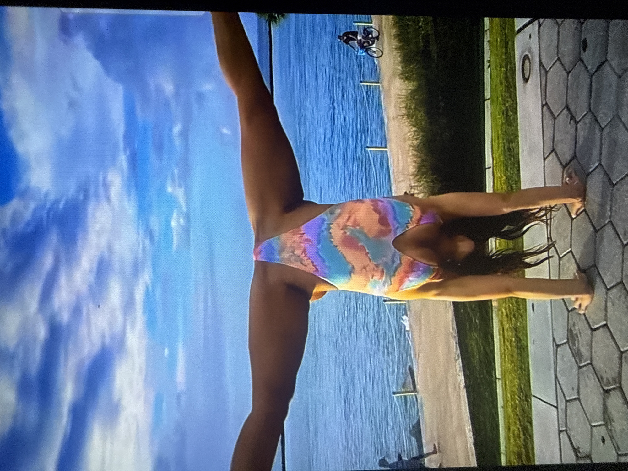 An outdoor scene in Varna, Bulgaria, depicts a female individual performing an inverted handstand with a wide leg split. The subject is positioned centrally, balancing on her hands on a grey hexagonal paved surface. She wears a multi-colored one-piece swimsuit, exhibiting pastel shades of blue, pink, orange, and purple in a tie-dye pattern. Her legs are extended horizontally in a full split, parallel to the ground. Her head is inverted towards the ground, with dark hair hanging downwards.
The immediate foreground consists of hexagonal grey pavers. Behind the subject, a strip of green grass is visible, followed by a light-colored concrete promenade. A body of water, identified as the Black Sea, extends behind the promenade, showing subtle wave patterns. Along the edge of the promenade adjacent to the water, a series of short white posts connected by a thin rope or barrier are present. In the background, on the promenade, a silhouetted individual is observed walking on the left side, while another individual rides a bicycle on the right side. The sky above is blue with scattered white clouds.