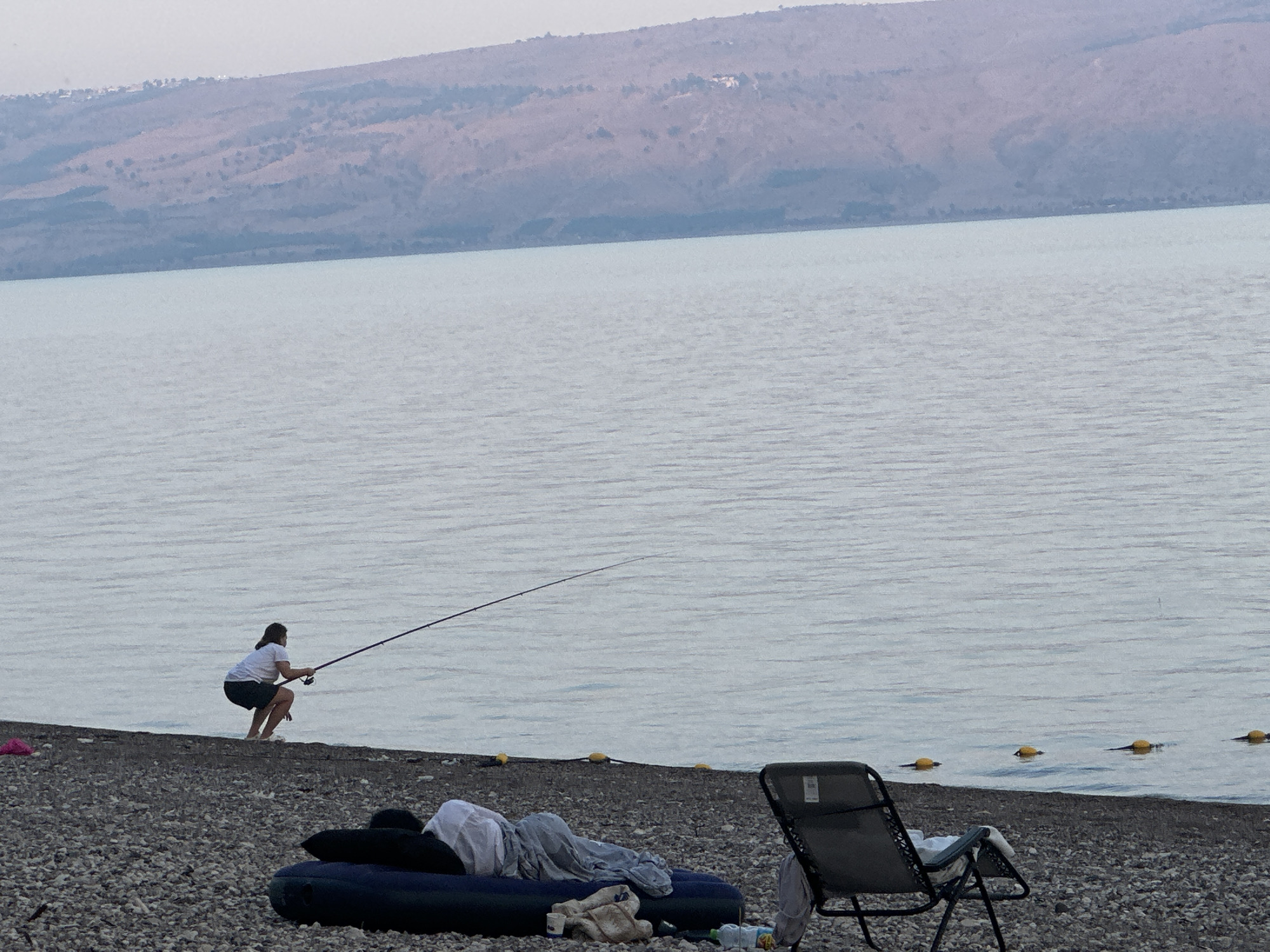 The image depicts a serene scene at the Sea of Galilee in Emek HaYarden Regional Council, Israel. In the foreground, a pebbly beach is dotted with camping gear, including an inflatable mattress with blankets and a folded lounge chair. A woman is actively fishing in the calm, light-blue water, holding a long fishing rod. In the background, a gently sloping mountain range, partially covered in vegetation, forms the horizon. Several yellow buoys are visible floating on the water's surface, marking a boundary. The overall atmosphere is tranquil, suggesting a peaceful outdoor activity.
