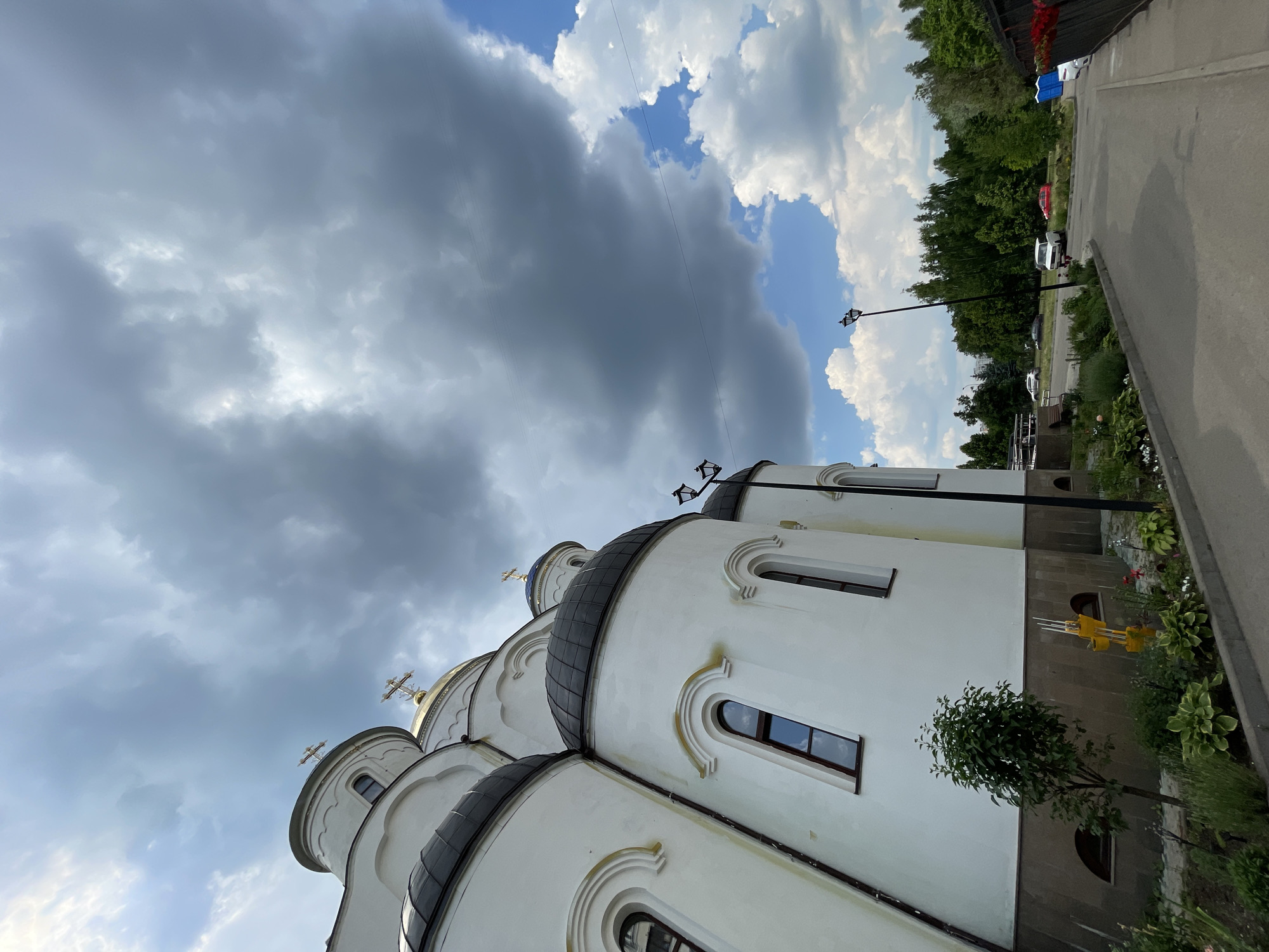 This is a low-angle, wide shot of the side of a white Orthodox church with black domes and golden crosses, under a cloudy, overcast sky with patches of blue. The church has several arched windows with dark frames. In the foreground, a paved walkway curves to the right, bordered by a small green tree, bushes, and flowers. Beyond the walkway, there are cars parked and trees in the background. The scene appears to be a sunny day with dramatic cloud formations. The location is in Moscow, Russia.