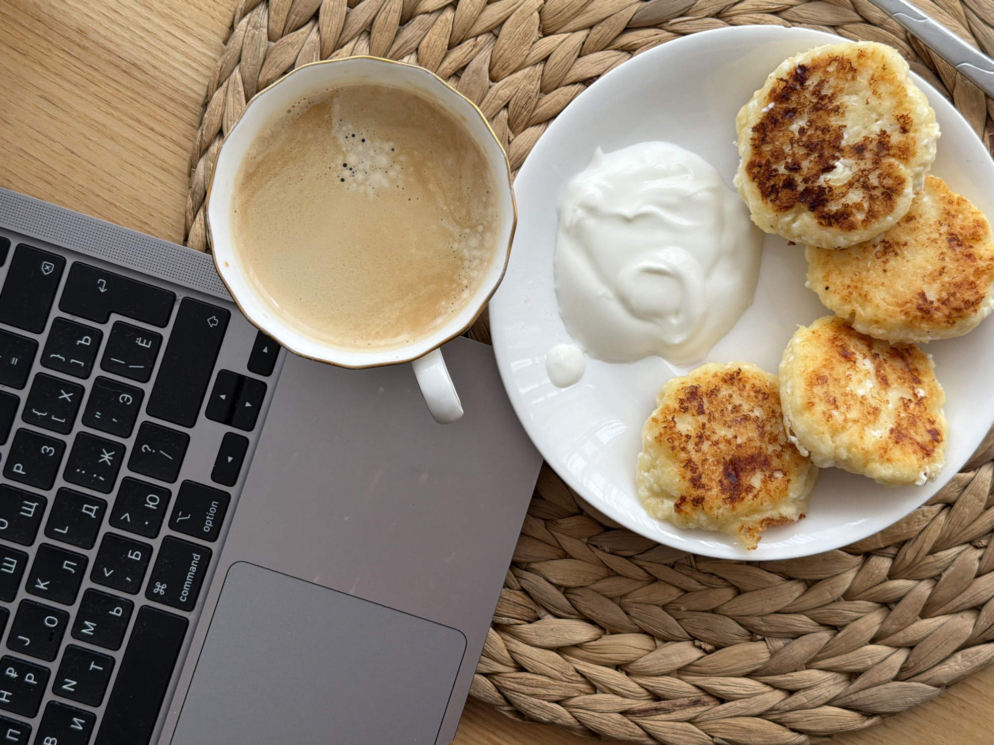 An overhead shot captures a breakfast or snack setup on a woven placemat, placed on a light wooden surface. The main subjects include a silver laptop, a white cup of coffee, and a white plate holding food.
On the plate, there are five golden-brown, pan-fried items, which appear to be syrniki (cottage cheese pancakes), accompanied by a dollop of thick white cream, likely sour cream or yogurt. A small portion of a silver utensil, possibly a fork or spoon, is visible on the right edge of the plate. Next to the plate, a white cup with an ornate gold rim contains a light brown beverage with a frothy top, indicative of coffee or a latte.
The laptop's keyboard features black keys with both Latin and Cyrillic characters, suggesting a Slavic language layout, consistent with the provided location context of Sofiivska Borshchahivka, Ukraine. Visible Cyrillic letters include Ж, Д, Л, О, П, И, Т, Б, Ю, Я, Ч, У, Ь, Э, Ы, Й, Ц, Ъ, Х, Ф, В, А, among others, alongside standard Latin keys and function keys like "command" and "option." The laptop's trackpad is also visible.
The woven placemat, made of natural fibers, adds a rustic texture to the scene. The overall arrangement suggests someone is taking a break or starting their day by combining work or leisure on the laptop with a meal. The bright, even lighting implies a daytime setting.