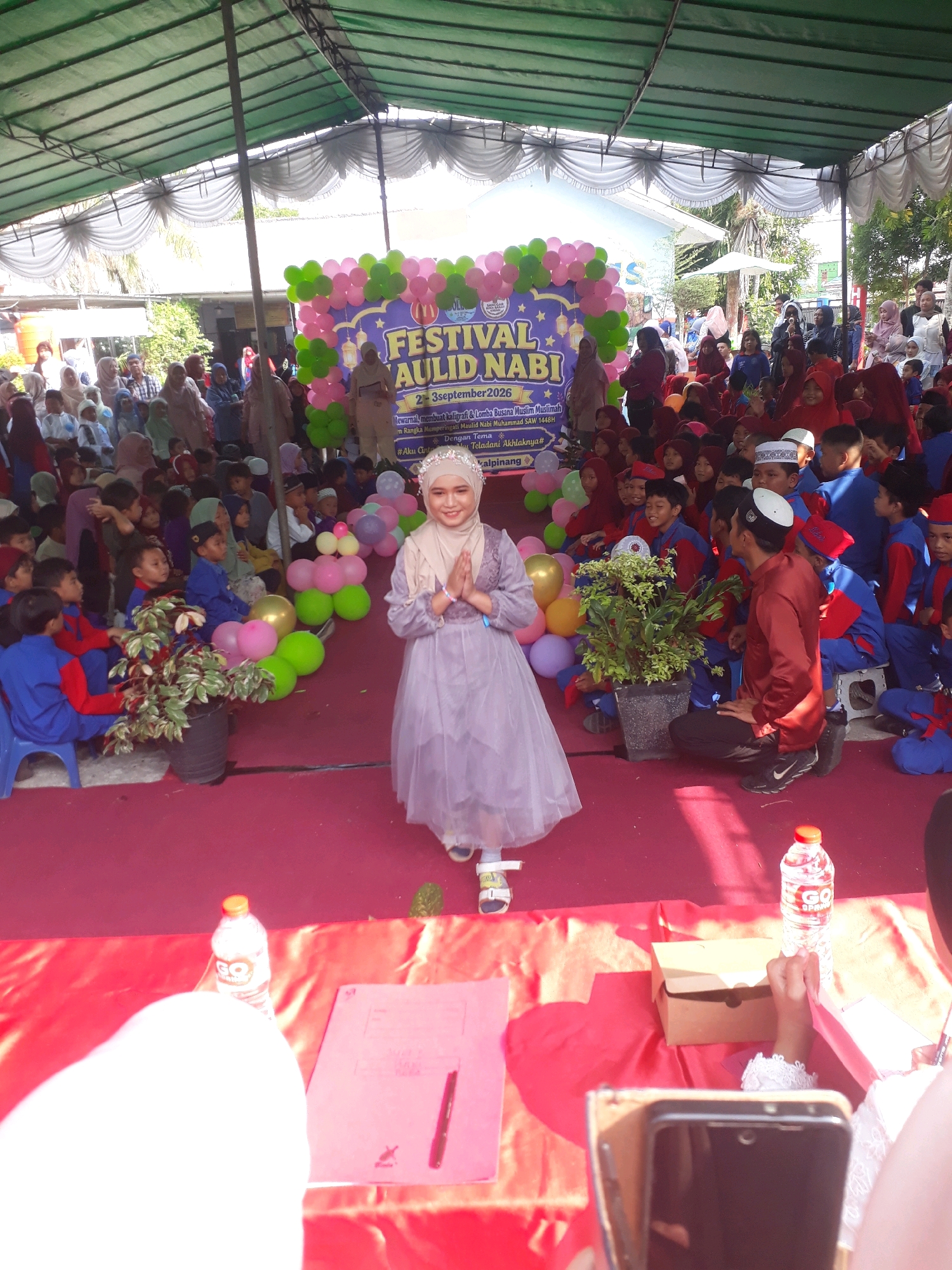 The image depicts an outdoor event, likely a festival, taking place under a green canopy structure. In the foreground, a young woman wearing a light purple dress and a hijab is posing with her hands clasped in front of her. She is standing on a red carpet. Behind her, a banner displays "FESTIVAL AULID NABBI" and other text in Indonesian, indicating a celebration related to the Prophet Muhammad. A crowd of children and adults are seated or standing around the area, many of them wearing traditional Islamic attire. Balloons in pink and green are arranged as a backdrop. To the right of the young woman, a potted plant adds a touch of greenery. On the red carpet, a bottle of "GO SPARKLE" water and a pink folder with a pen are visible, along with a cardboard box. The environment suggests a public gathering, possibly at a school or community center, given the presence of many children. The signage on the banner suggests the event is in Pangkalpinang, Indonesia, and is related to a religious celebration.