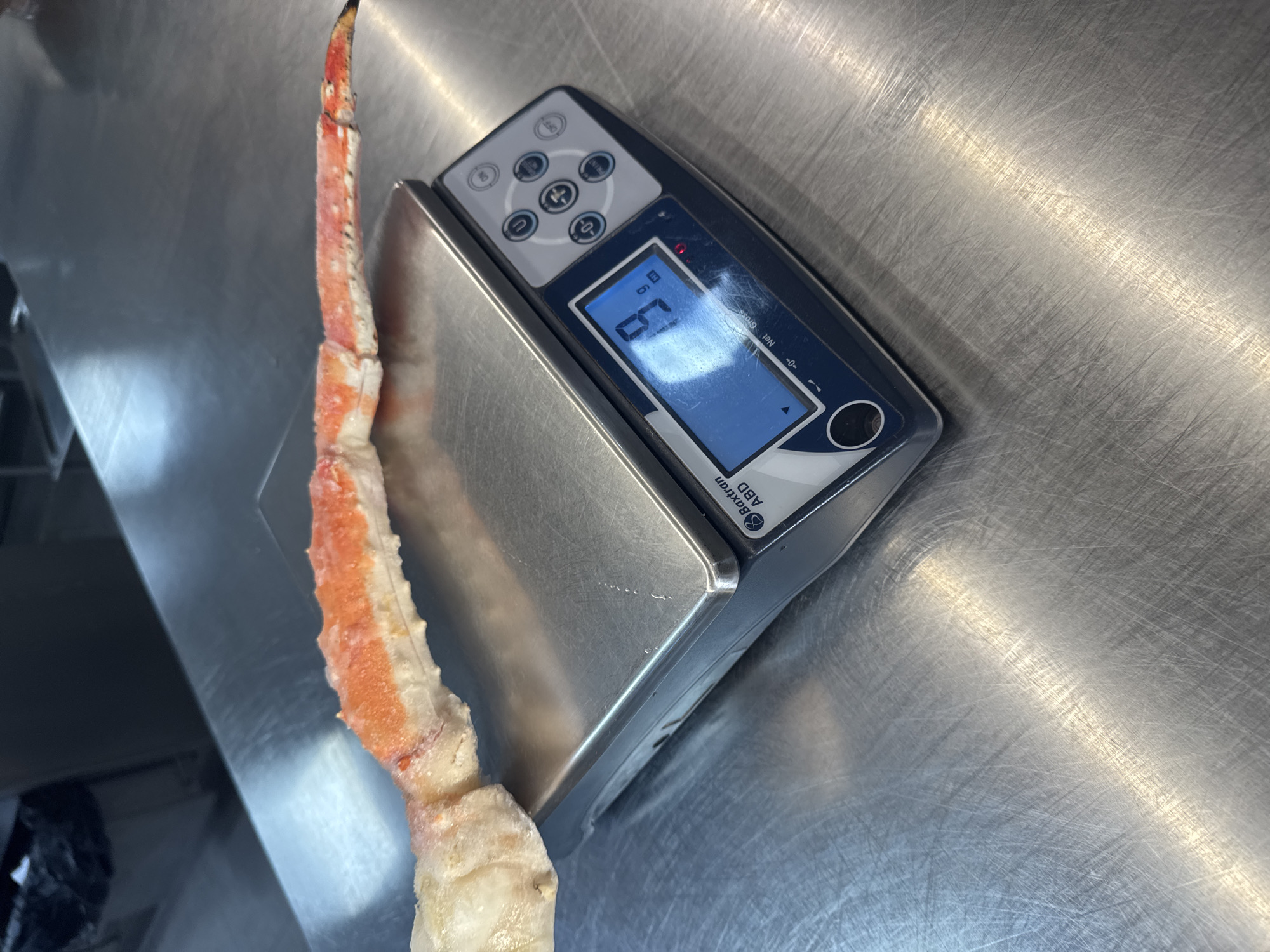 A crab leg is being weighed on a stainless steel digital scale. The scale is placed on a stainless steel surface. The display on the scale reads "69". The brand name "Baxktron ABD" is visible on the scale. The scene appears to be in a commercial kitchen or food processing environment. There is no indication of this image being in Sofia, Bulgaria.