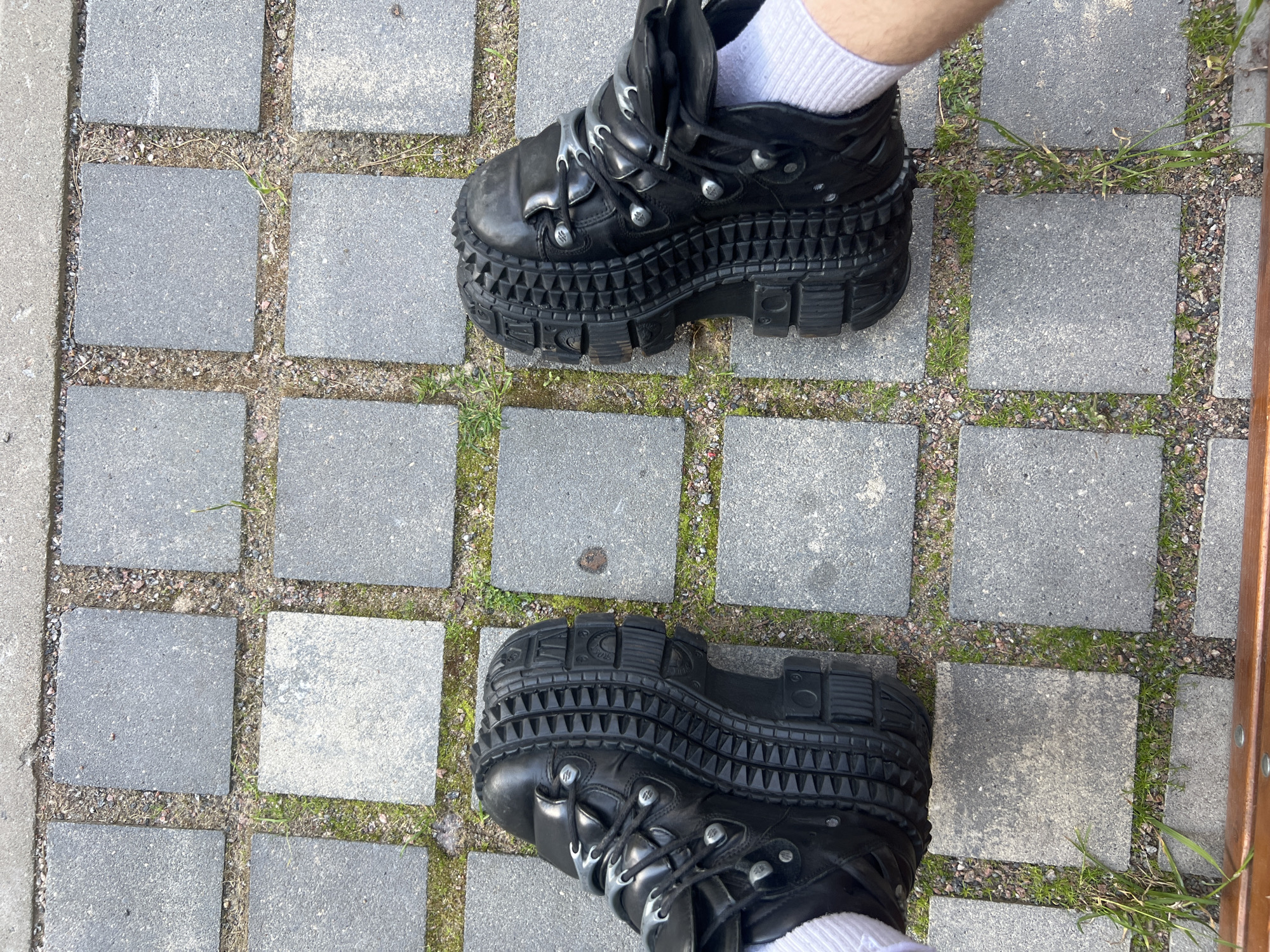 Two feet, clad in white ribbed socks and prominent black platform boots, are centered on a paved outdoor surface, viewed from a downward perspective. The boots feature multiple straps, metallic eyelets, and a heavily textured sole with a distinct, almost spiky, ridged pattern on the sides. The sole of the right boot clearly displays the embossed text "NEW ROCK." The boots appear somewhat worn, indicating use.
The ground beneath is composed of square, grey concrete or stone pavers, which are laid with gaps filled with small gravel and sparse green weeds or grass. To the left, there is a narrow strip of concrete, and on the right, a segment of a wooden structure, possibly a bench leg or railing, with metallic accents is visible. The lighting is bright and even, suggesting it is daytime. The overall scene captures a moment of standing or walking on a casual outdoor path in Saint Petersburg, Russia.