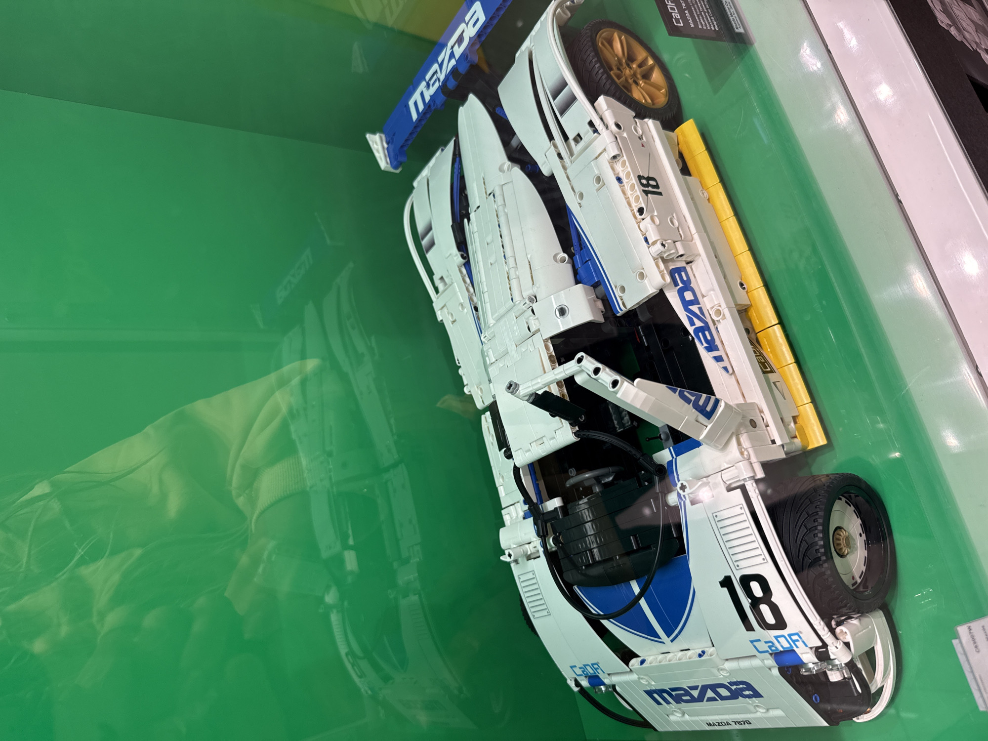 The image shows a detailed Lego Technic model of a white and blue Mazda 787B race car with gold wheels. The car is on display behind a green, transparent surface, likely glass, which creates reflections of the surrounding environment. The Lego model is white with blue stripes and the word "MAZDA" printed in blue on the side. The number "18" is prominently displayed on the side as well. A small information plaque with "CaDF" and "Mazda 787B" is visible in the lower right corner. The background is a solid green color with faint, out-of-focus reflections of what appear to be people and possibly other displays. The scene is indoors, suggested by the controlled lighting and the reflective display case. There is no indication of the specific location in Yaroslavl, Russia, as no identifying landmarks or signs are visible.