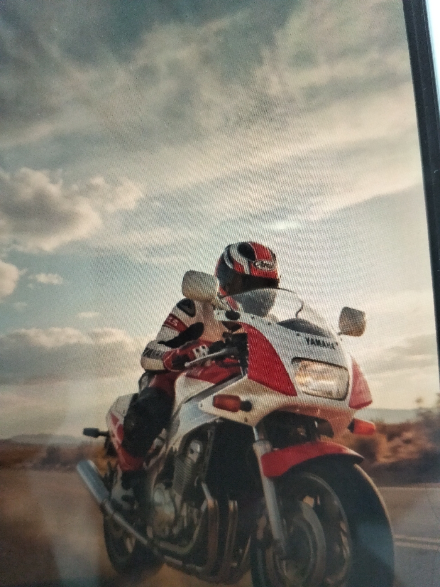 A motorcyclist wearing a red and white racing suit and a red and white helmet is riding a red and white motorcycle. The motorcycle has the word "YAMAHA" written on it. The motorcyclist is in motion, as evidenced by the dust being kicked up around the front wheel. The background appears to be a desert landscape with mountains in the distance and a cloudy sky above. There is no visible indication of this scene being located in the city of Payagaboh, Indonesia.
